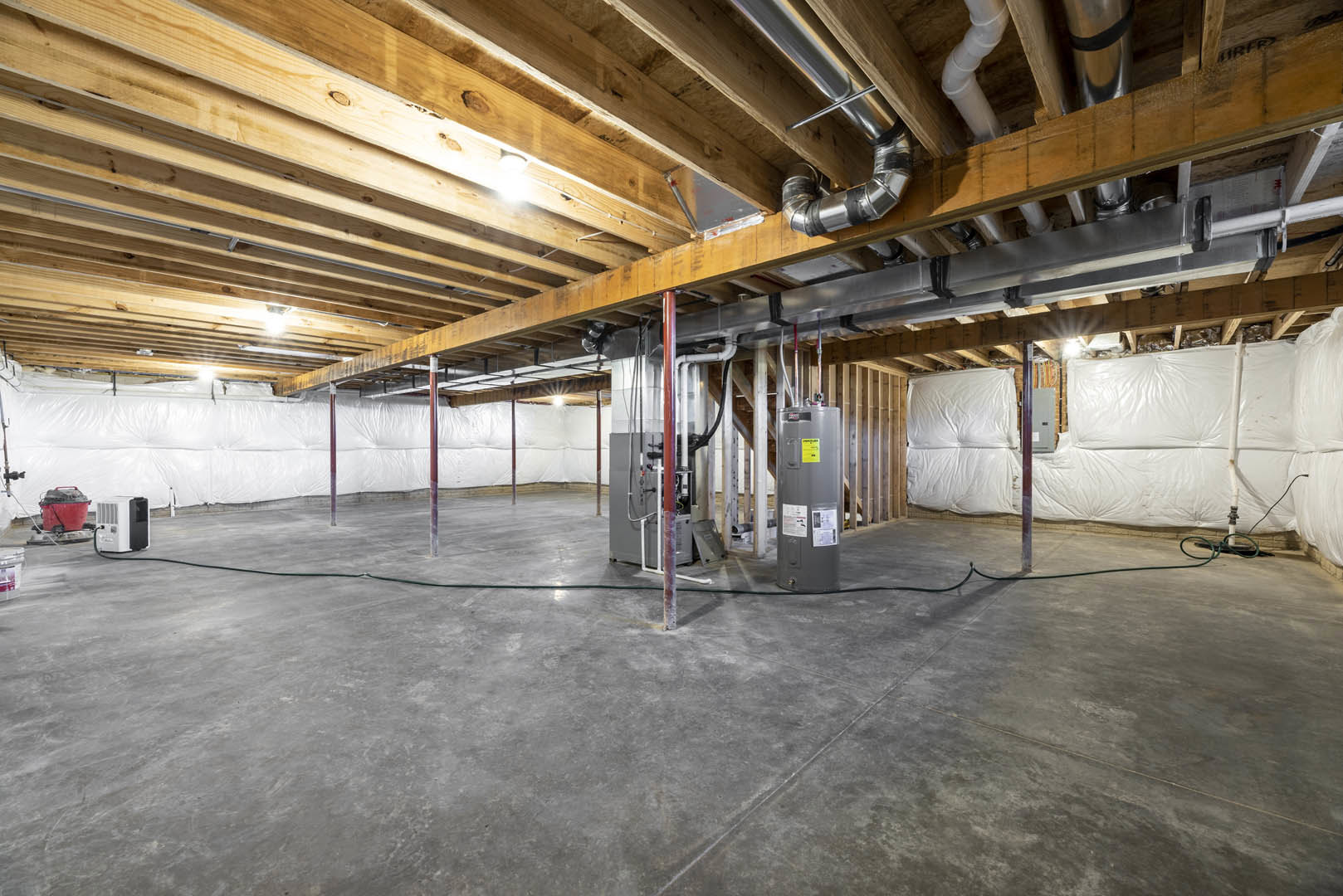 Basement room with exposed steel beams, white plastic insulation, concrete floor, large grey cylinder, red bucket, and white utility box.