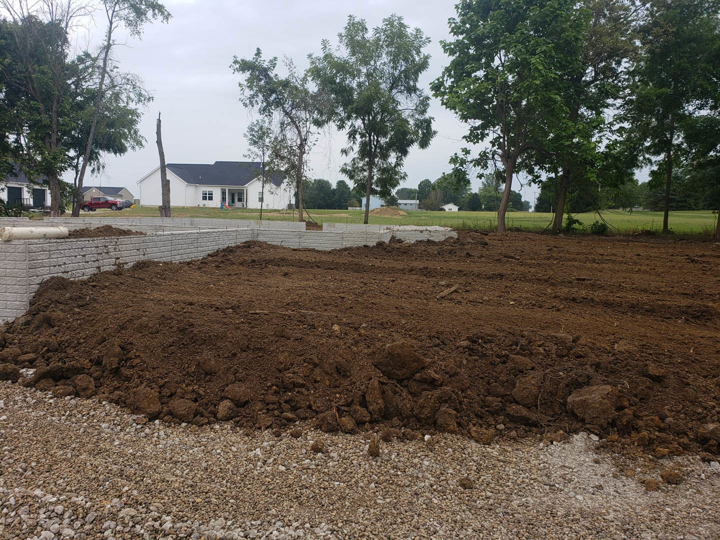 White house with black roof behind large dirt pile in grassy yard, surrounded by green-leaved trees under blue sky