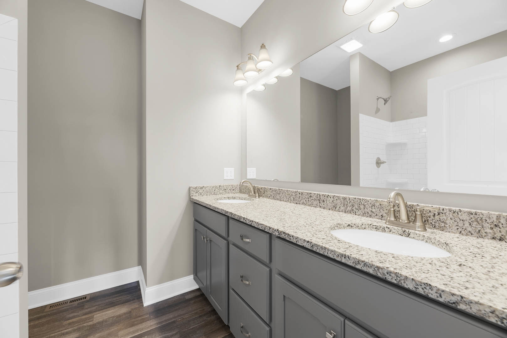 Bathroom with expansive mirror above gray stone countertops, white tile backsplash, modern chrome faucet, wood flooring with white trim, and white door against white walls.