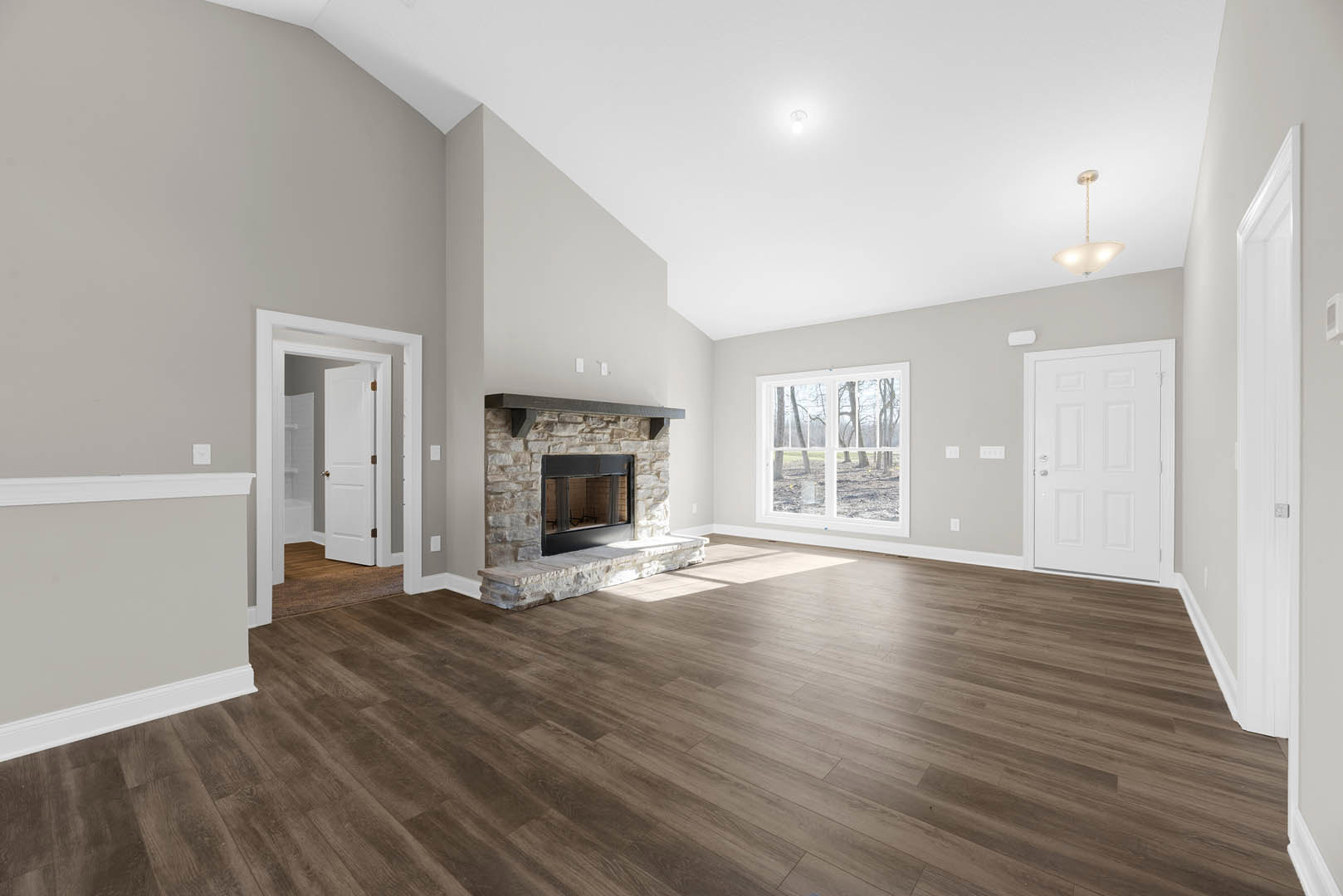 Spacious living room featuring hardwood floors, central stone fireplace, white walls, large window with tree views, and a white door with silver handle