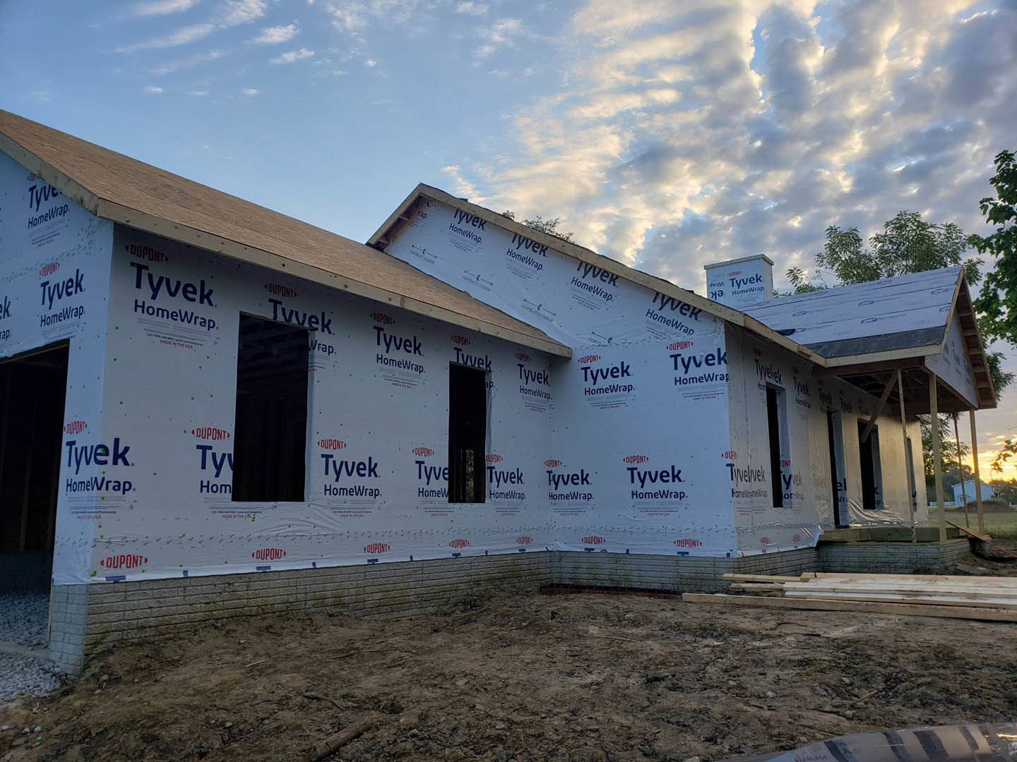 Framed custom home under construction with exposed wood beams, unfinished brick walls, black front door with red paint, dirt ground, white interior wall visible through window