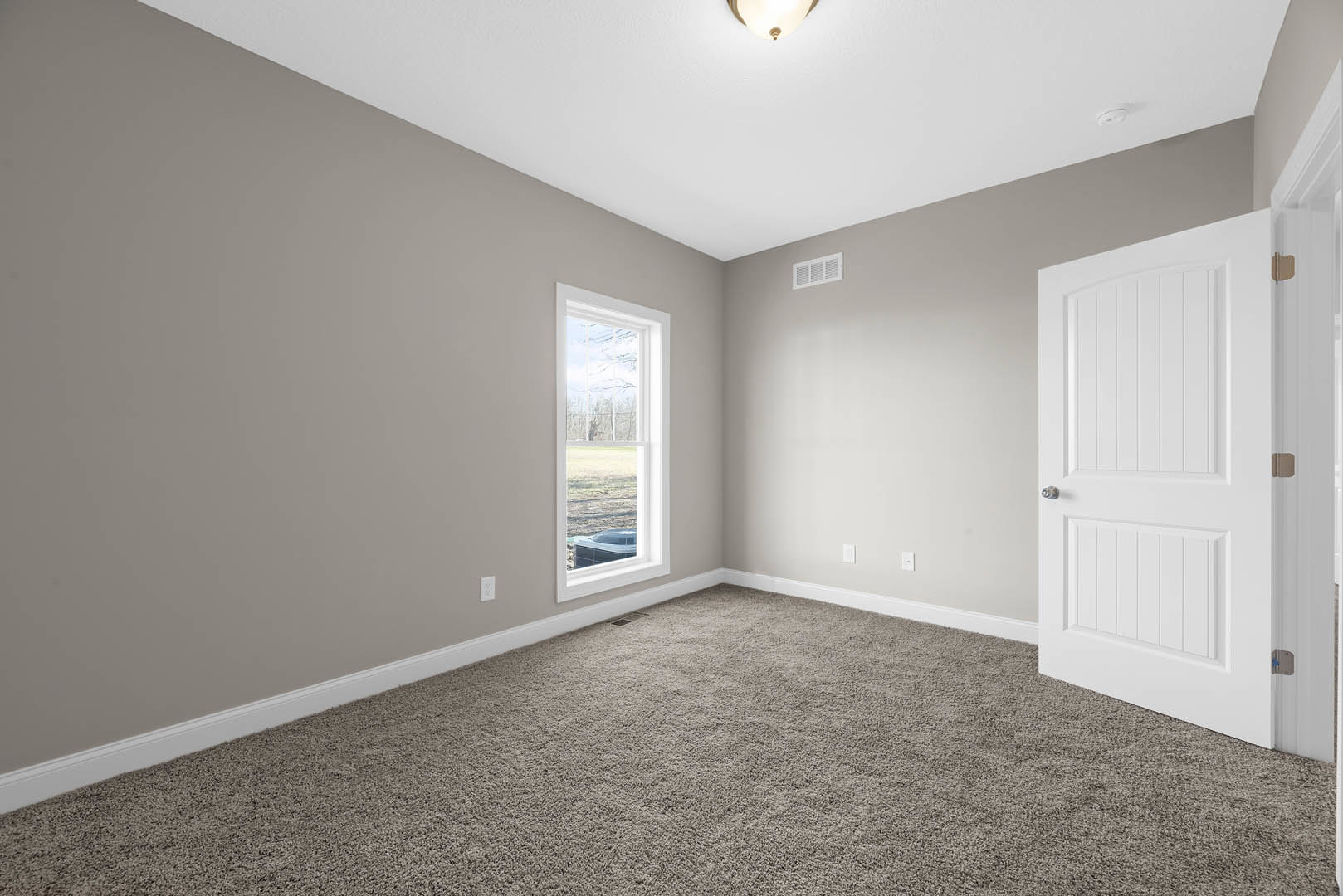 Neutral-toned carpeted room with white paneled door, wall vent, large window overlooking grassy yard and trees, white baseboards, and smooth plaster walls