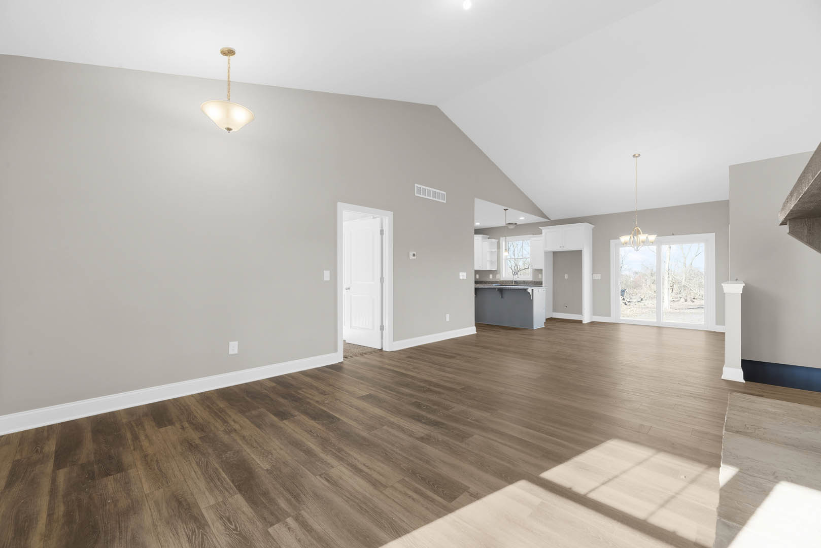 Spacious room with light wood flooring, white walls, ceiling light fixture, white door with silver handles, kitchen countertop, and window overlooking outdoors