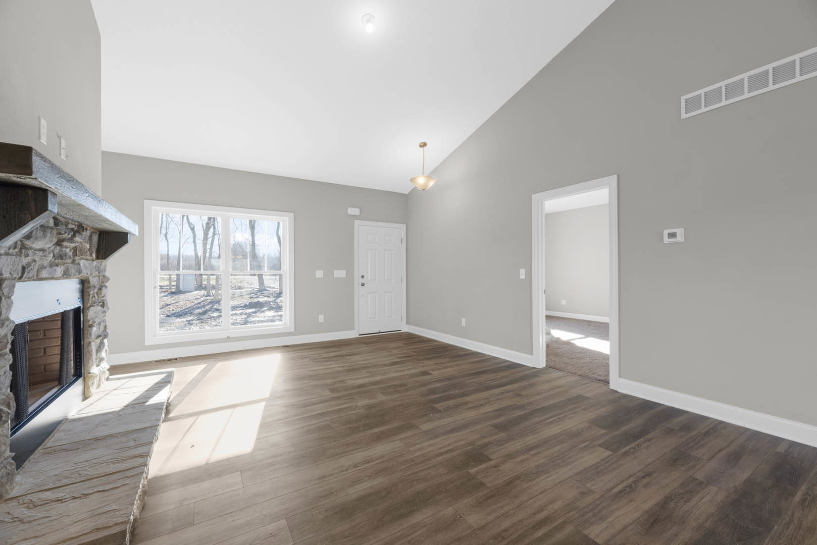 Wood flooring in a room with a white door featuring a silver handle, stone fireplace, tall floor lamp, and window overlooking trees