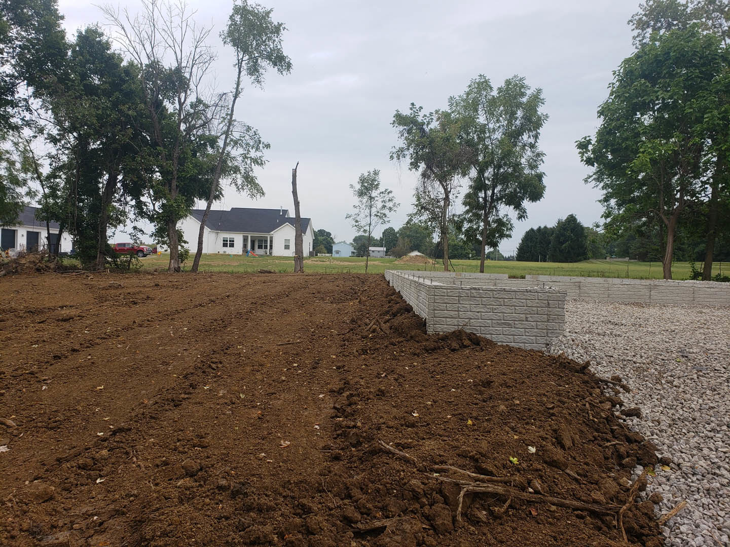 Dirt field bordered by mature trees, white house with brick wall and playground visible in the background under partly cloudy sky