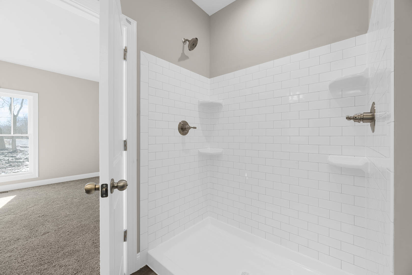 White tile bathroom featuring a walk-in shower with two built-in shelves, chrome showerhead and faucet, window overlooking a tree, and a door with metal hardware.