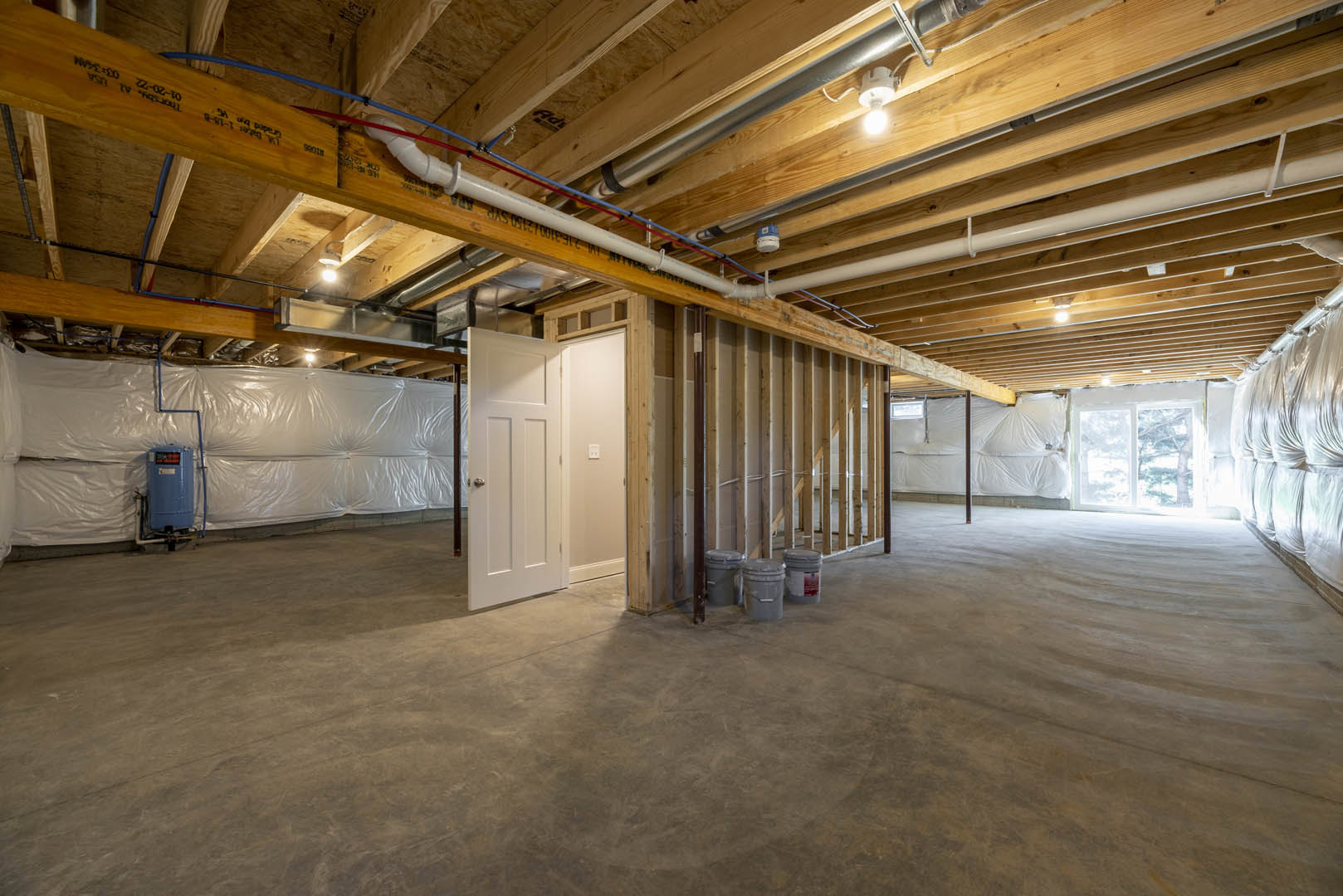 Basement room with exposed wood beam ceiling, visible pipes, blue water tank with attached pipe, concrete floor, grey bucket, and close-up of a door.