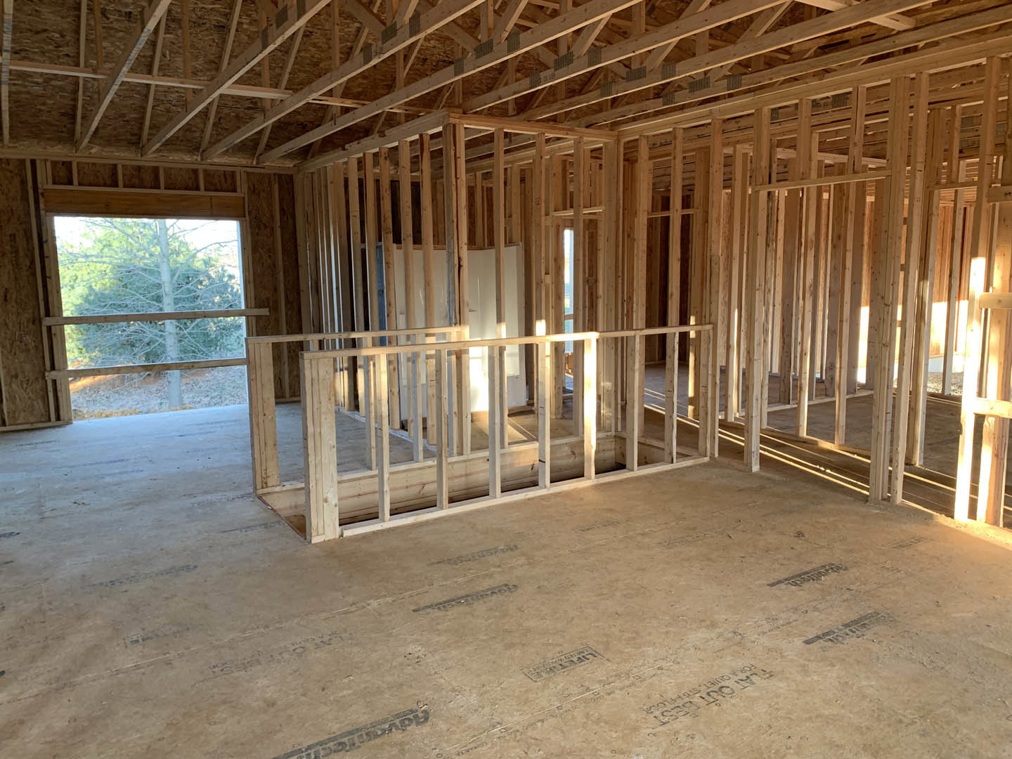 Exposed wood framing and beams inside a partially constructed home, unfinished floors, ceiling, and window with tree visible outside