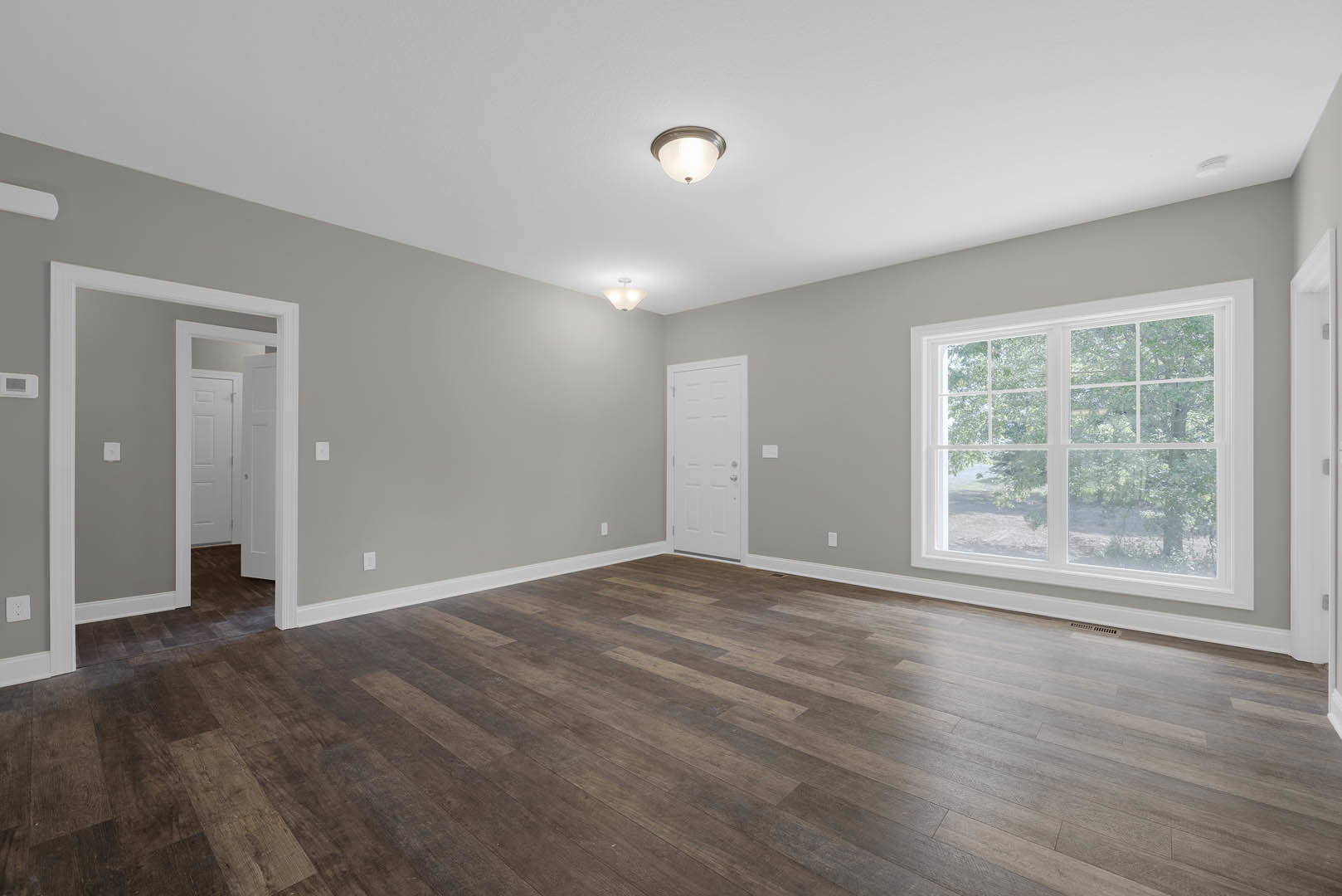 Wood flooring in a bright room with a large window overlooking trees, white walls, a white door with a silver handle, and a ceiling light fixture