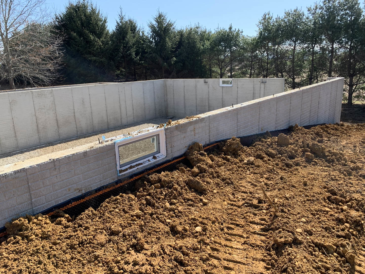 Concrete exterior wall with rectangular window, bare tree and row of trees in background, pile of soil next to foundation, overcast sky above.