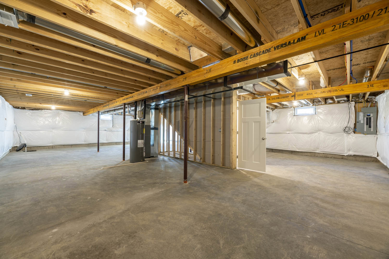 Room with exposed wooden ceiling beams, white walls, concrete floor featuring a central pipe, white door with silver knob, single ceiling light bulb, and white plastic wall