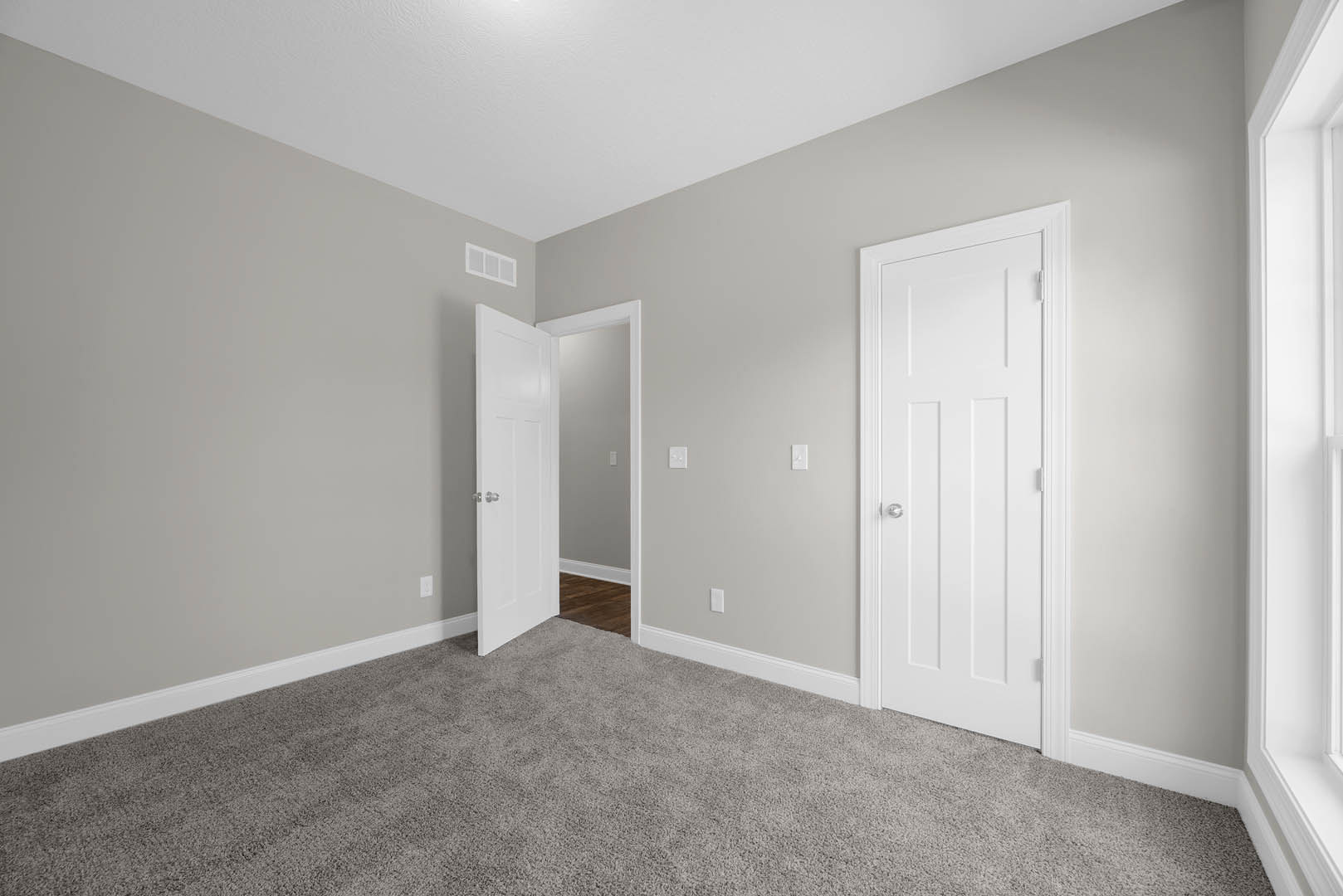 Carpeted room with two white doors featuring silver knobs, white baseboards, and partial view of wood laminate flooring