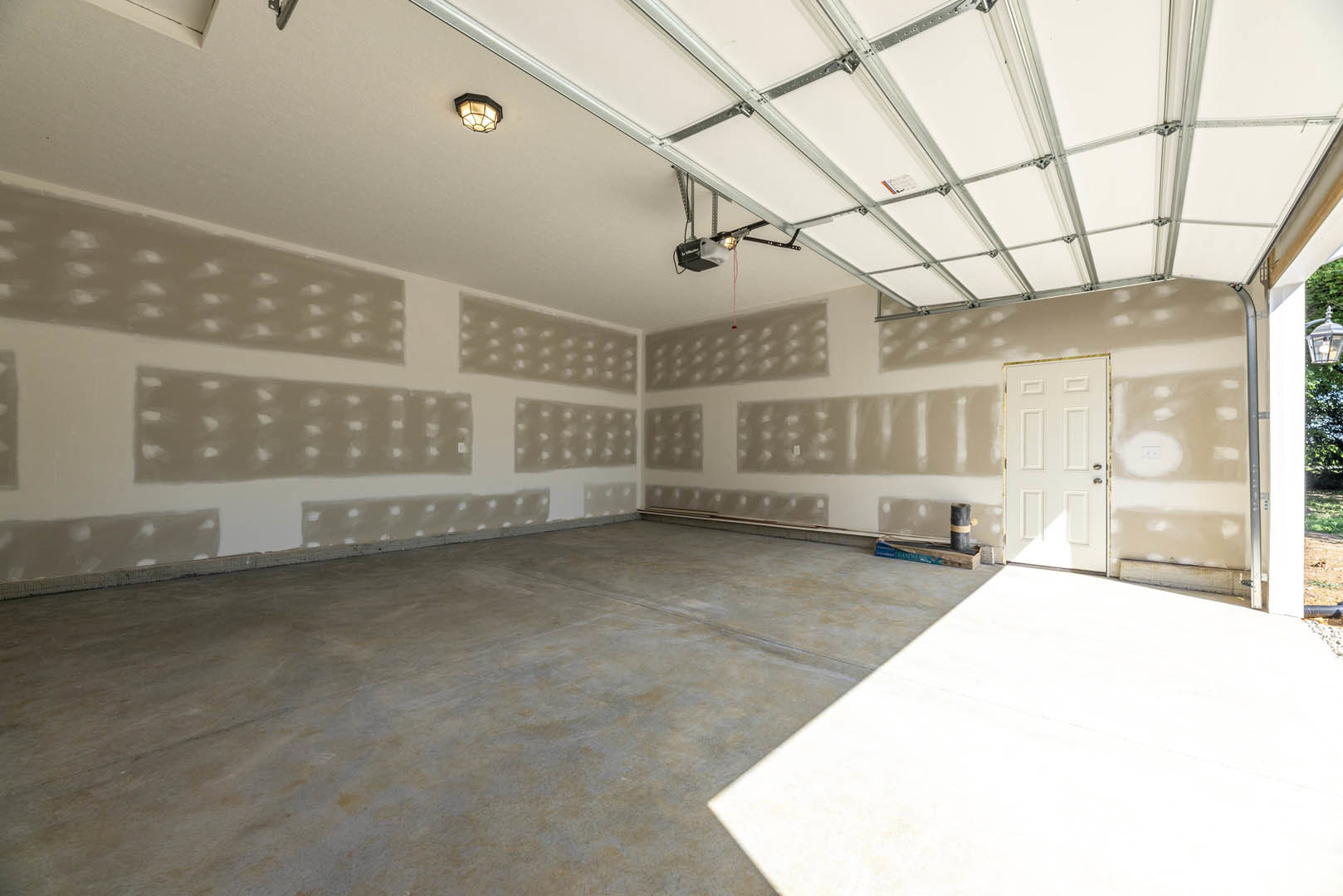 White-walled room featuring a white door with a silver handle, white concrete floor, white ceiling with exposed metal framing, ceiling light fixture, and a garage door with a
