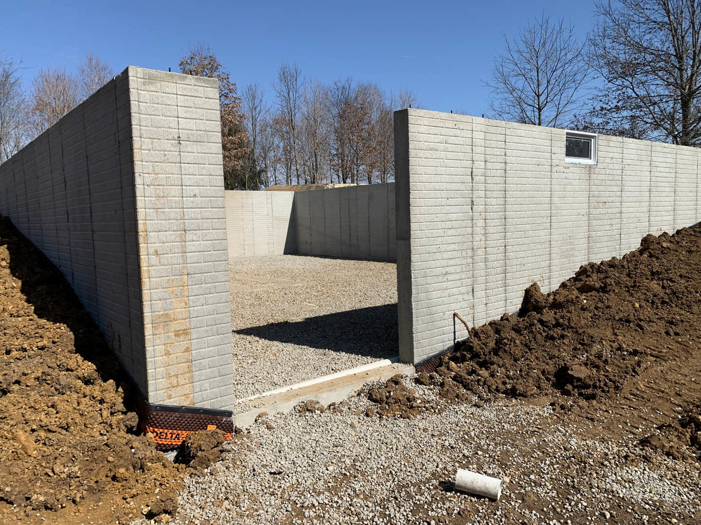 Concrete foundation wall with rectangular opening, exposed dirt and trees in background, white pipe lying on ground, partial view of brick wall and black and orange construction