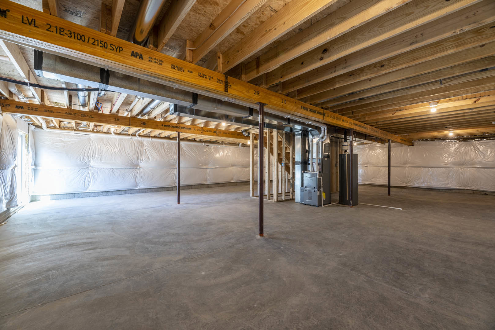 Exposed wooden ceiling beams and metal pipes above a concrete floor, white plastic bags stacked near a wall draped with a white sheet, unfinished basement interior