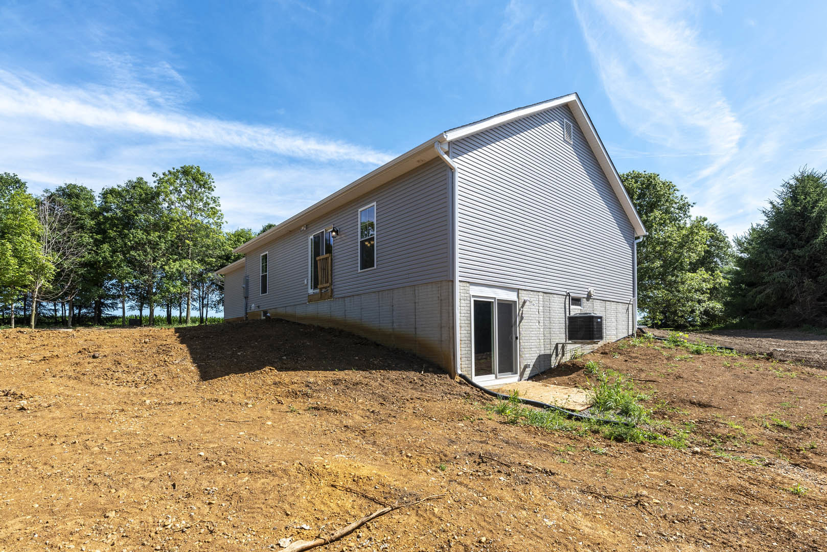 Modern cottage-style home with light siding and dark roof, situated on a grassy hill with a dirt slope, surrounded by mature trees under a clear blue sky