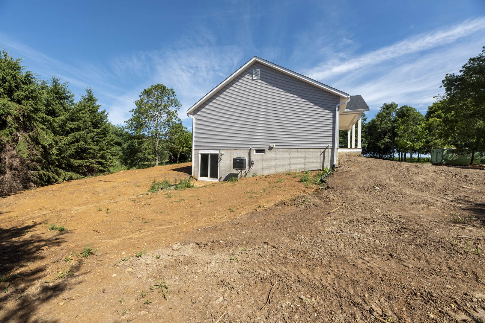 Modern brick home with black-framed windows and dark roof, situated atop a dirt hill with grassy patches, tall green tree nearby, and cloudy sky overhead