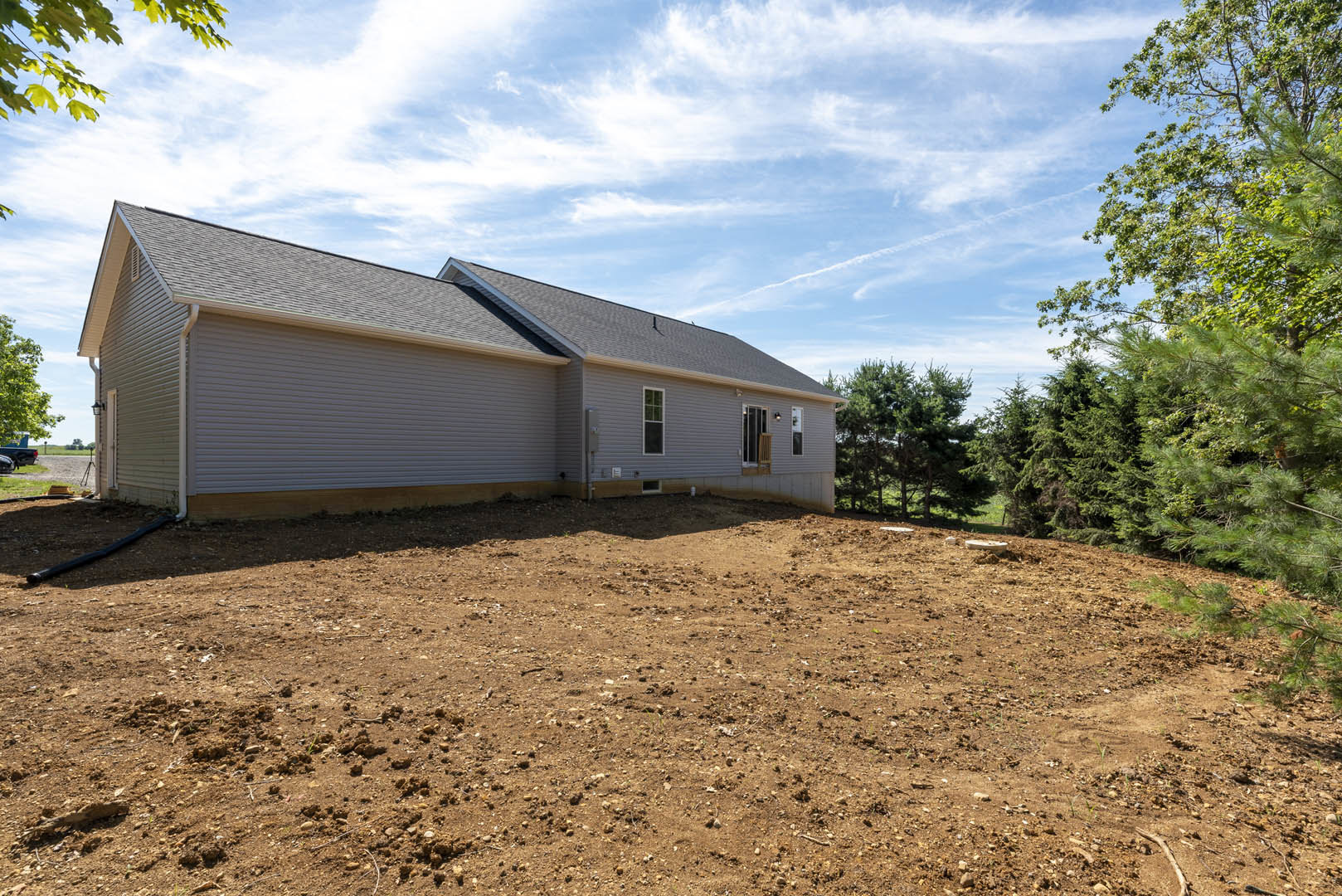 Two-story custom home with grey roof, surrounded by dirt lot and scattered trees, black pipe lying on ground, blue sky with clouds overhead