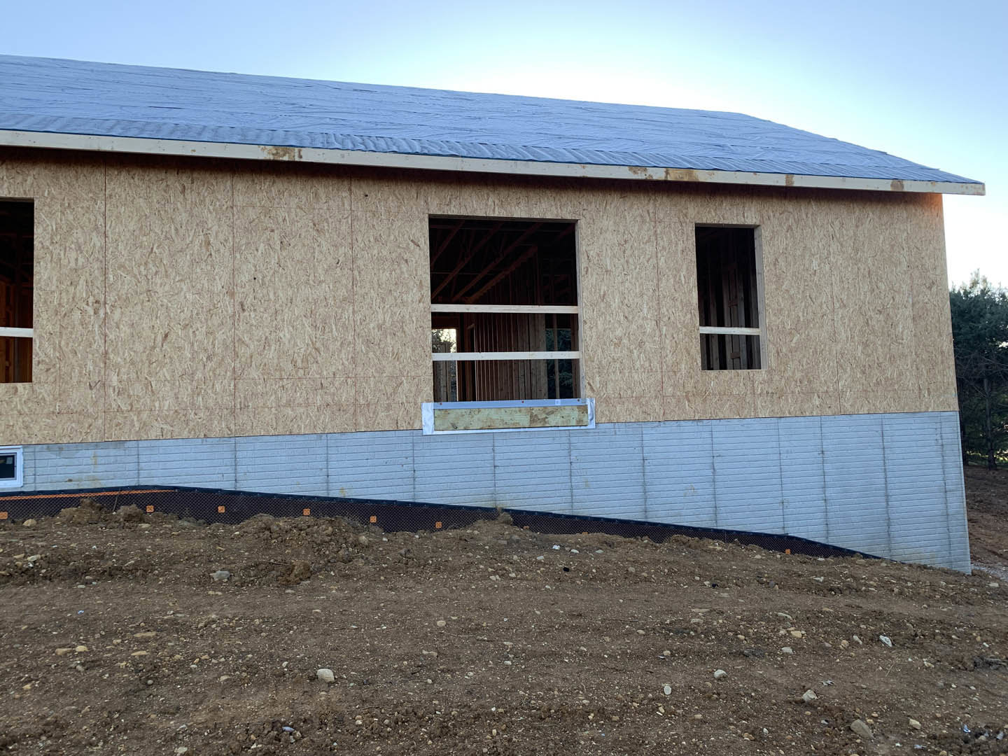 Wood-framed custom home under construction with blue metal roof, exposed beams, unfinished siding, and dirt hill bordered by a fence