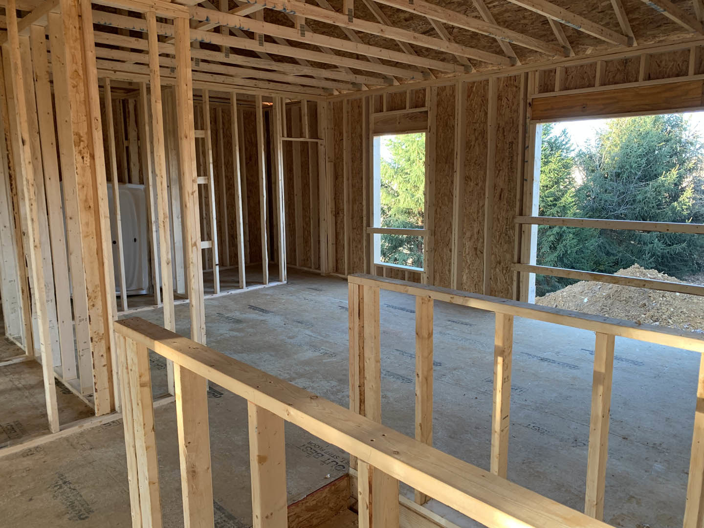 Exposed wooden beams and framing structure of a house under construction, large window opening with view of green trees, white door with black hardware visible
