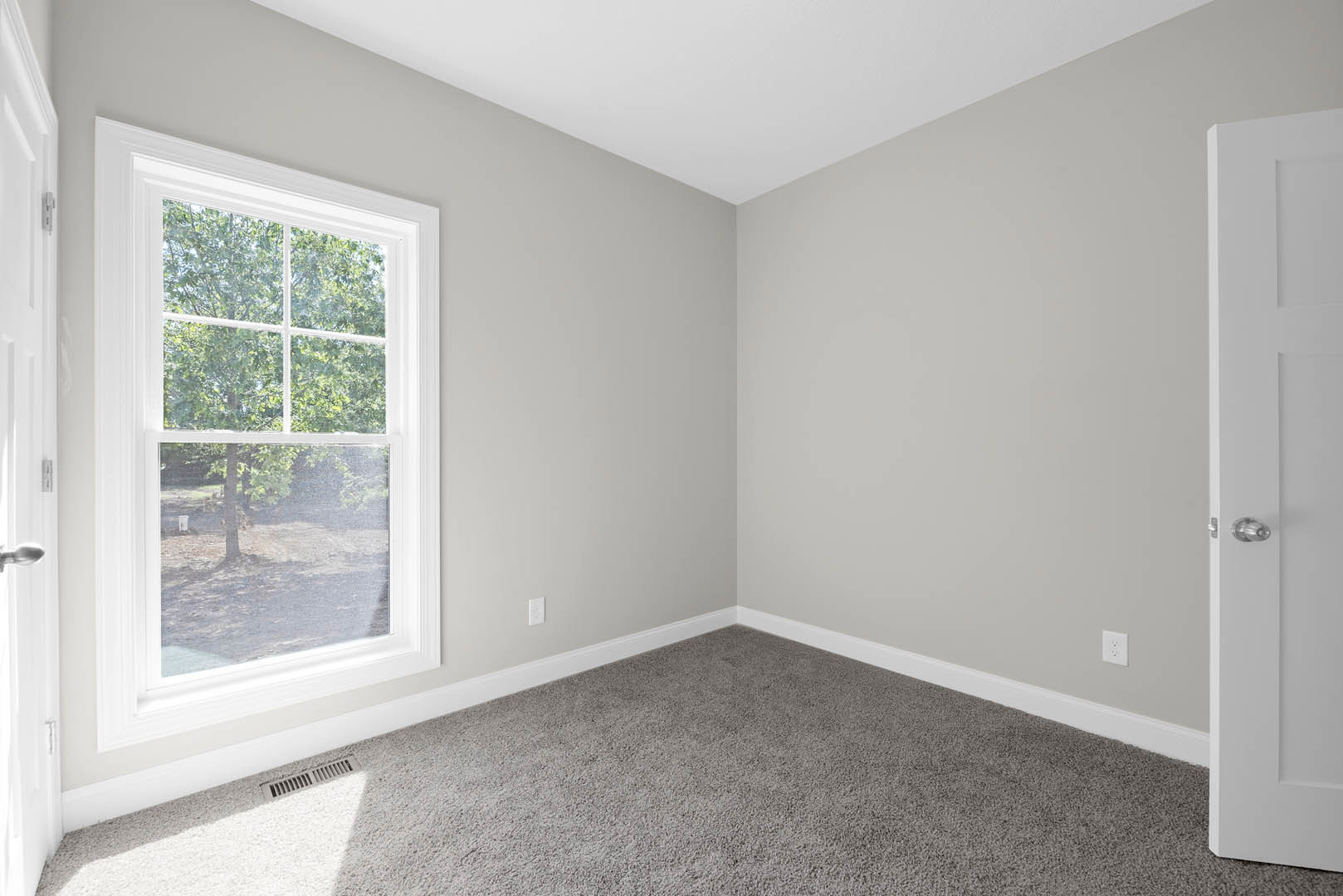 Bedroom with beige carpet, white walls, large window overlooking a tree, white door with silver knob, and floor vent near the baseboard