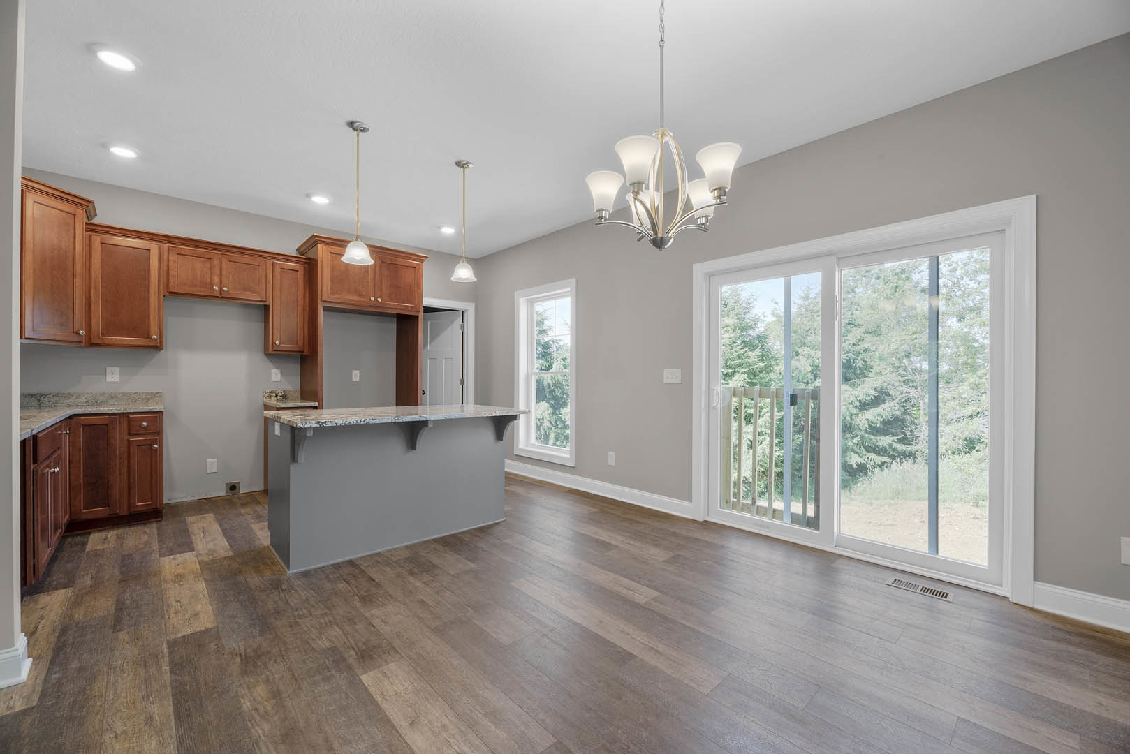 Open kitchen and dining area with wood flooring, marble countertop, white cabinetry, chandelier, sliding glass door, and large window overlooking trees.