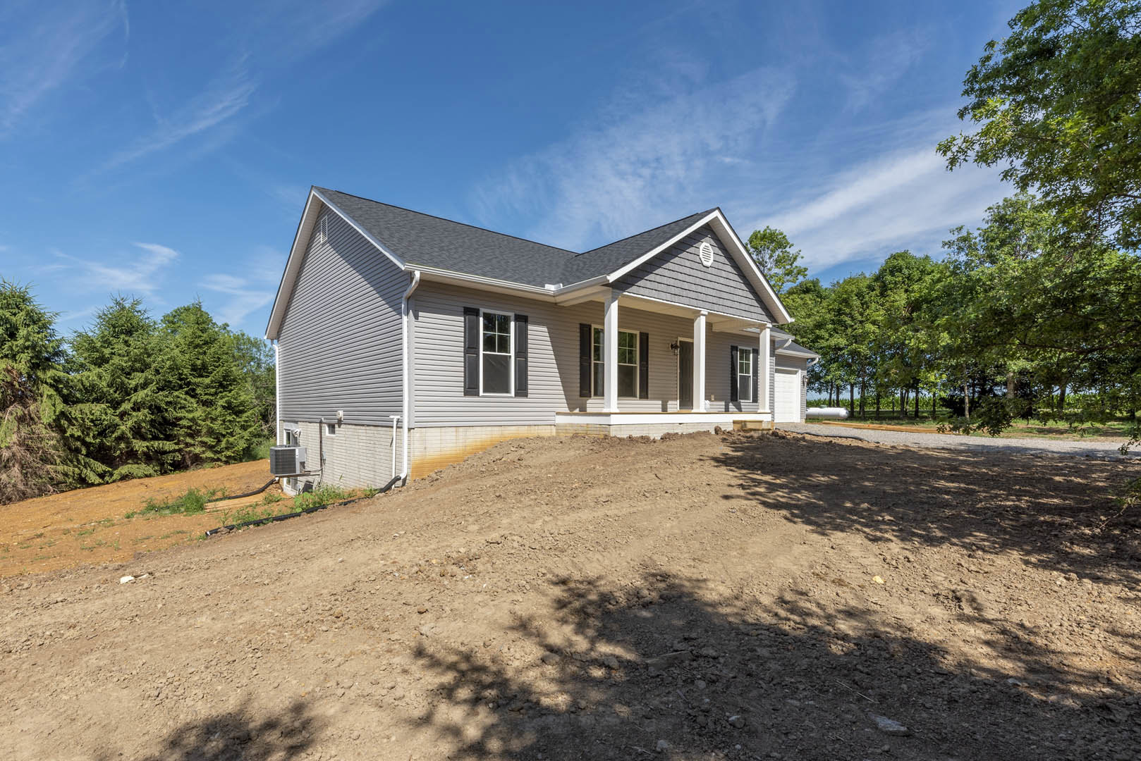Two-story home with white-framed windows, covered porch, air conditioner unit, dirt hill in front, and mature trees under a blue sky