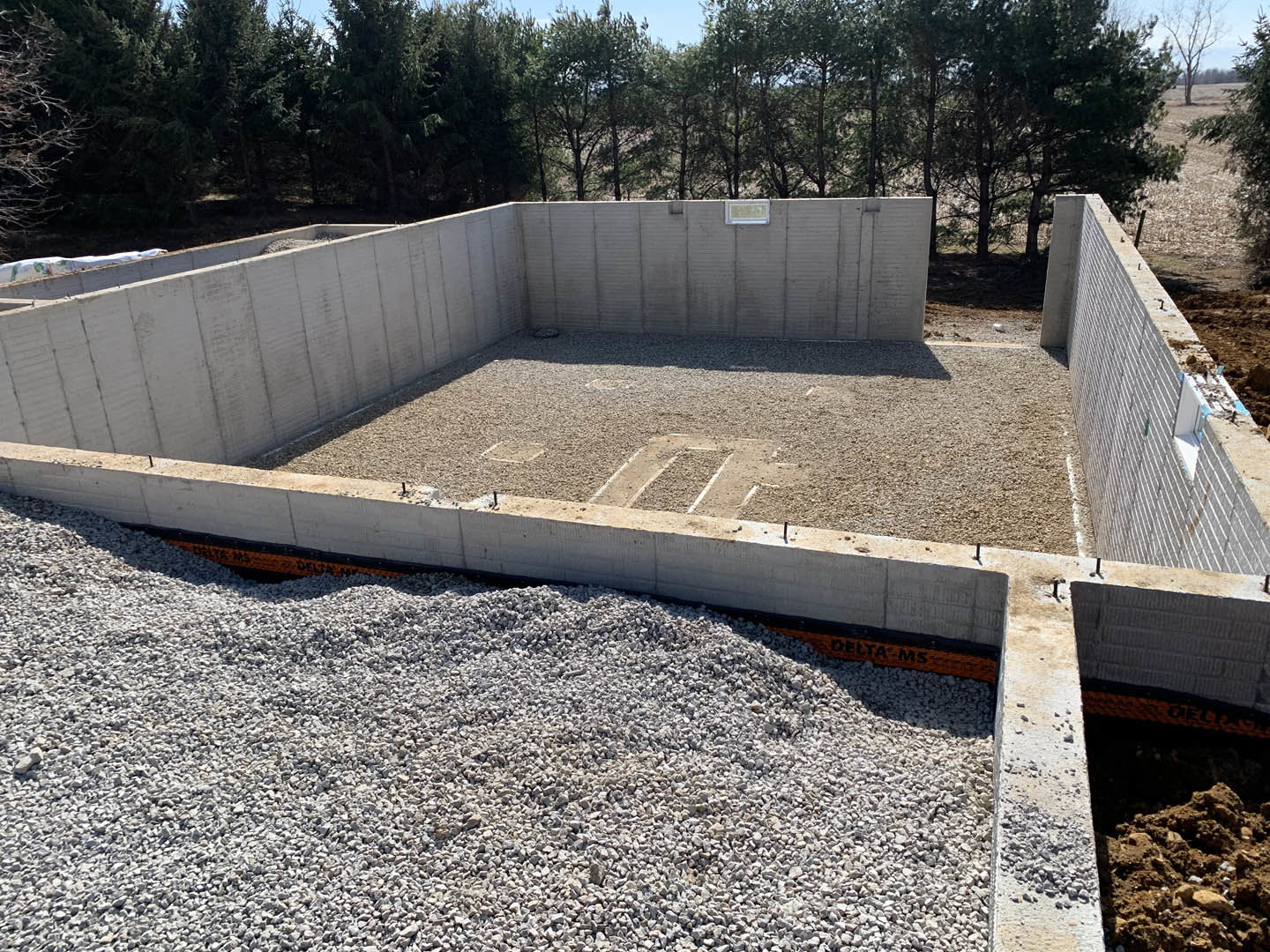 Concrete foundation surrounded by gravel and a large gravel pile, with trees and sky in the background, dirt and rocks scattered near a concrete wall casting a shadow