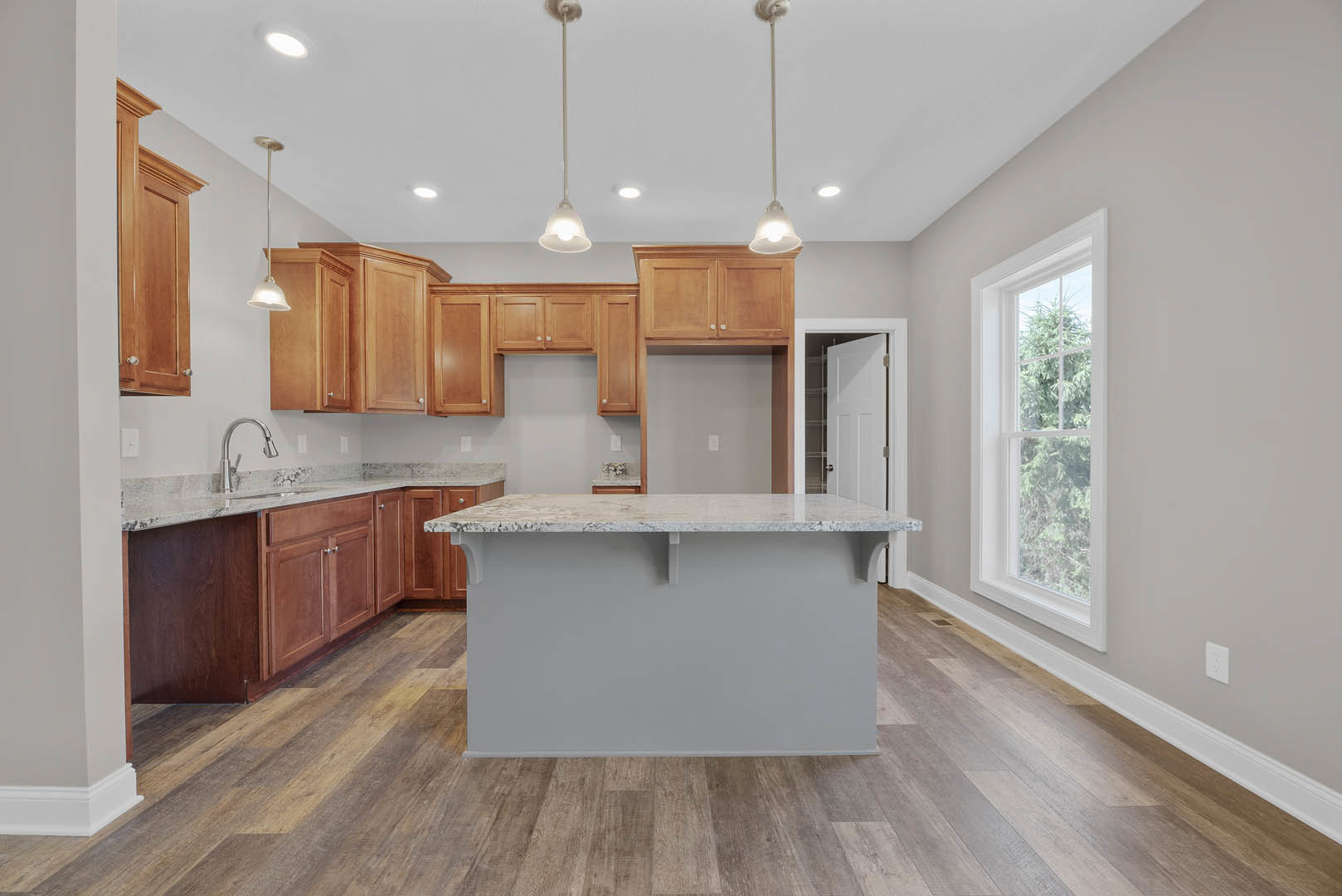 Kitchen with central island, wooden cabinets, tile flooring, stone countertop, stainless steel faucet, pendant light fixture, white walls, window overlooking trees.