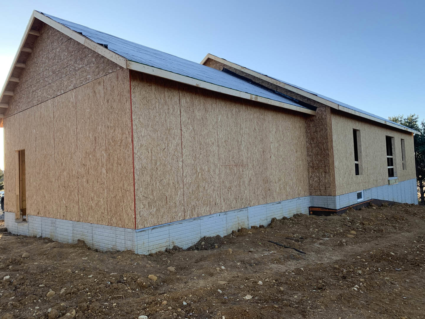 Wood-framed house under construction with exposed beams, unfinished walls, dirt and rocks in foreground, clear blue sky overhead