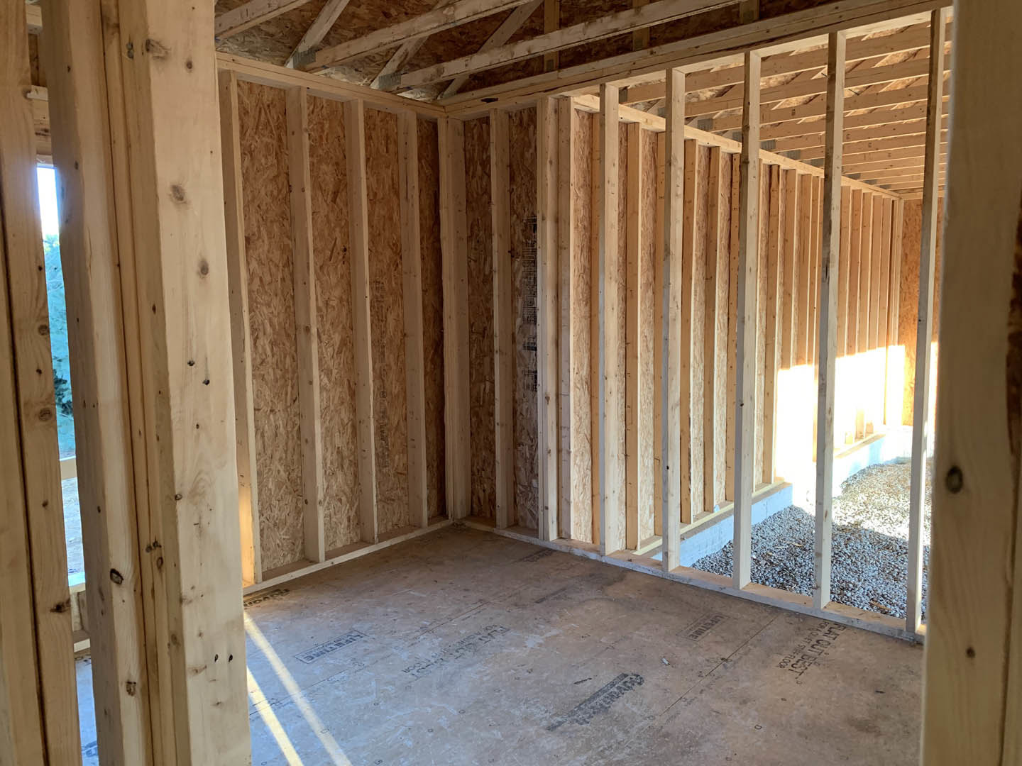 Wood-framed room with exposed ceiling beams, unfinished walls, and a window; construction materials visible on floor.