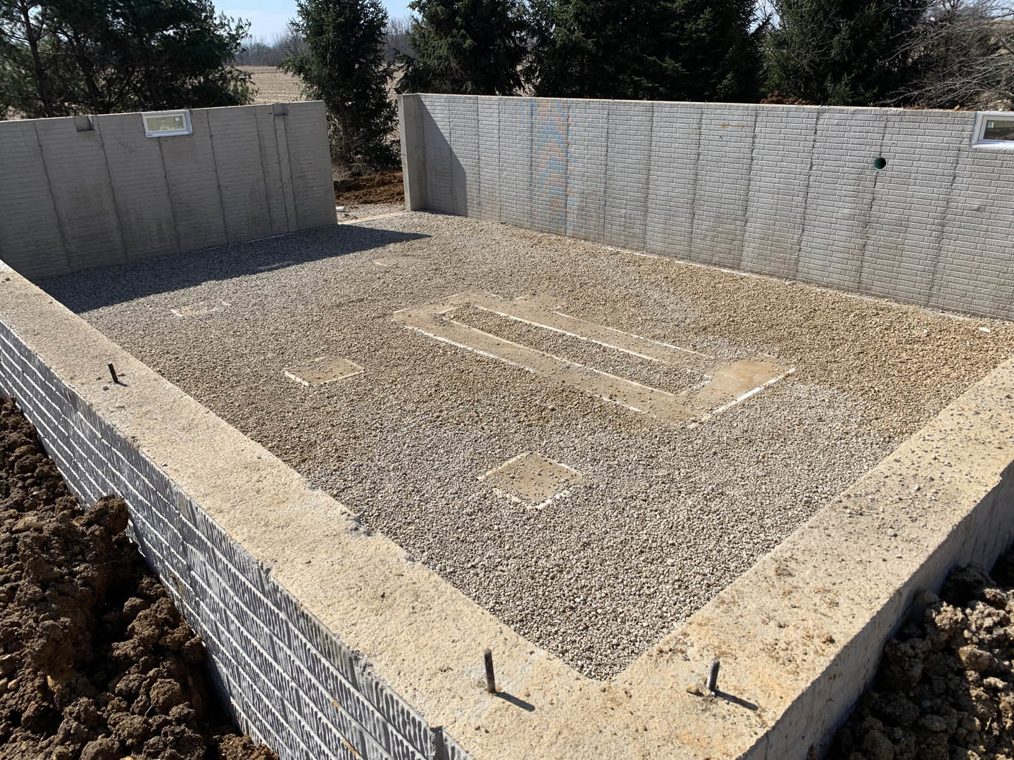Concrete foundation with gravel base, grey concrete wall featuring a small window, brick wall and trees visible in background.