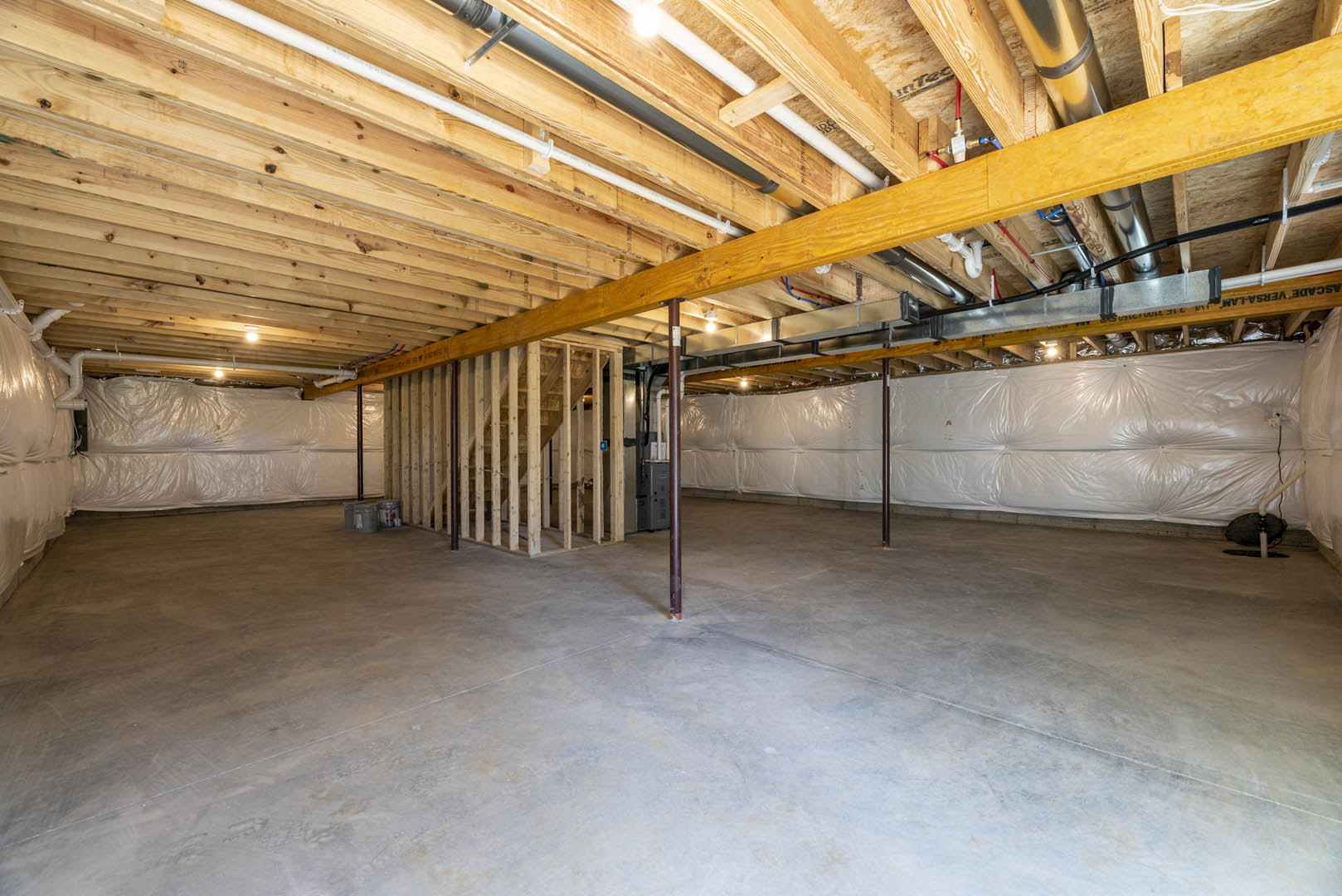 Basement interior with exposed wood beams and ceiling, concrete floor, central metal pole, white plastic wall covering, visible pipes along wooden ceiling.