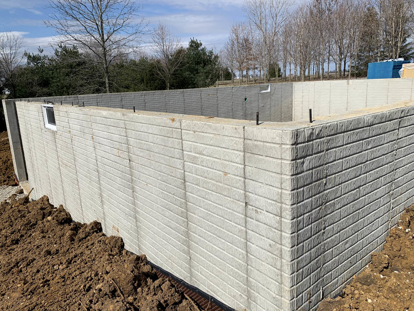 Concrete exterior wall with rectangular ground opening, exposed dirt, leafless tree, and blue sky in background