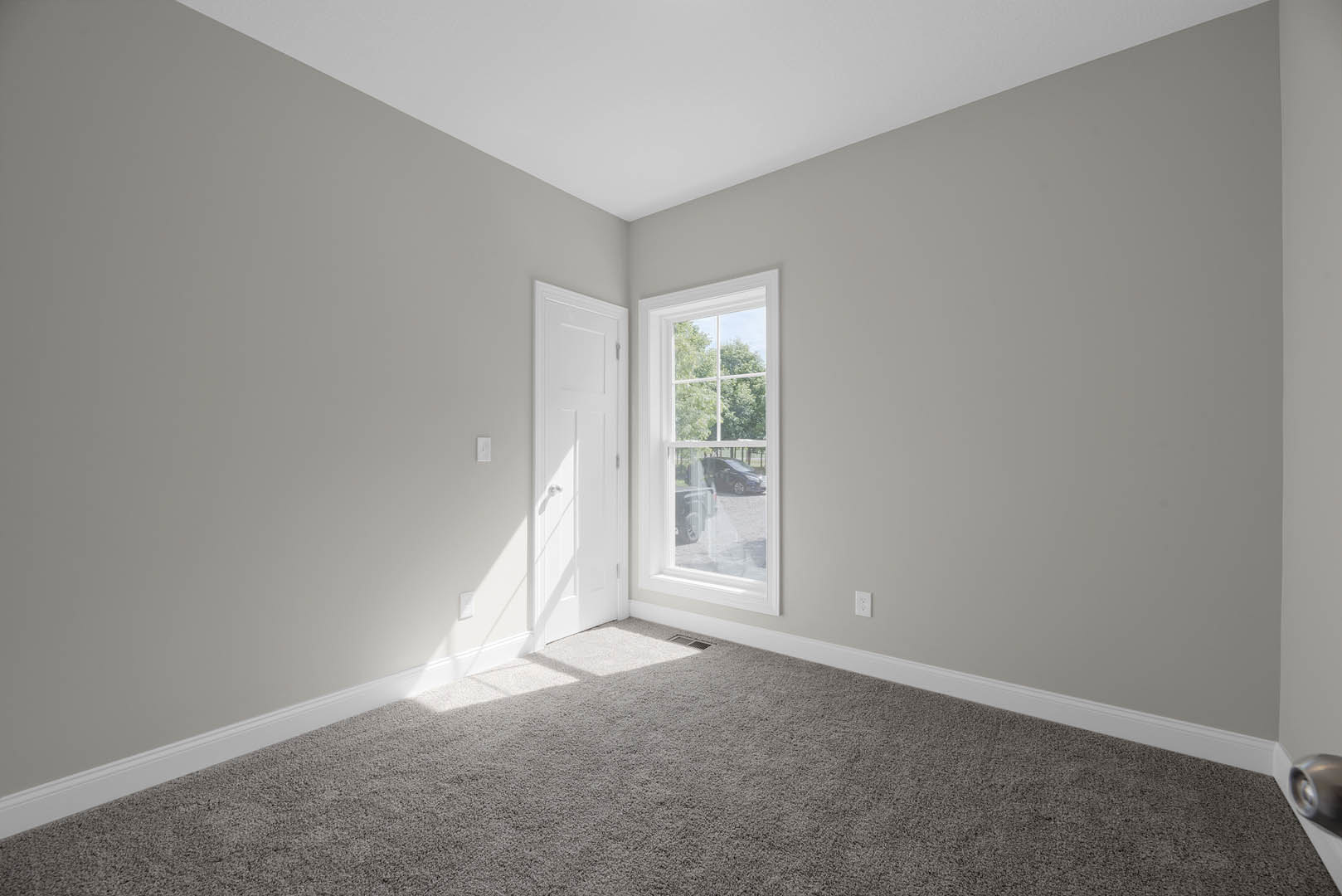 Neutral-toned carpeted room with white paneled door, window overlooking parked car, light-colored walls, and simple baseboard molding