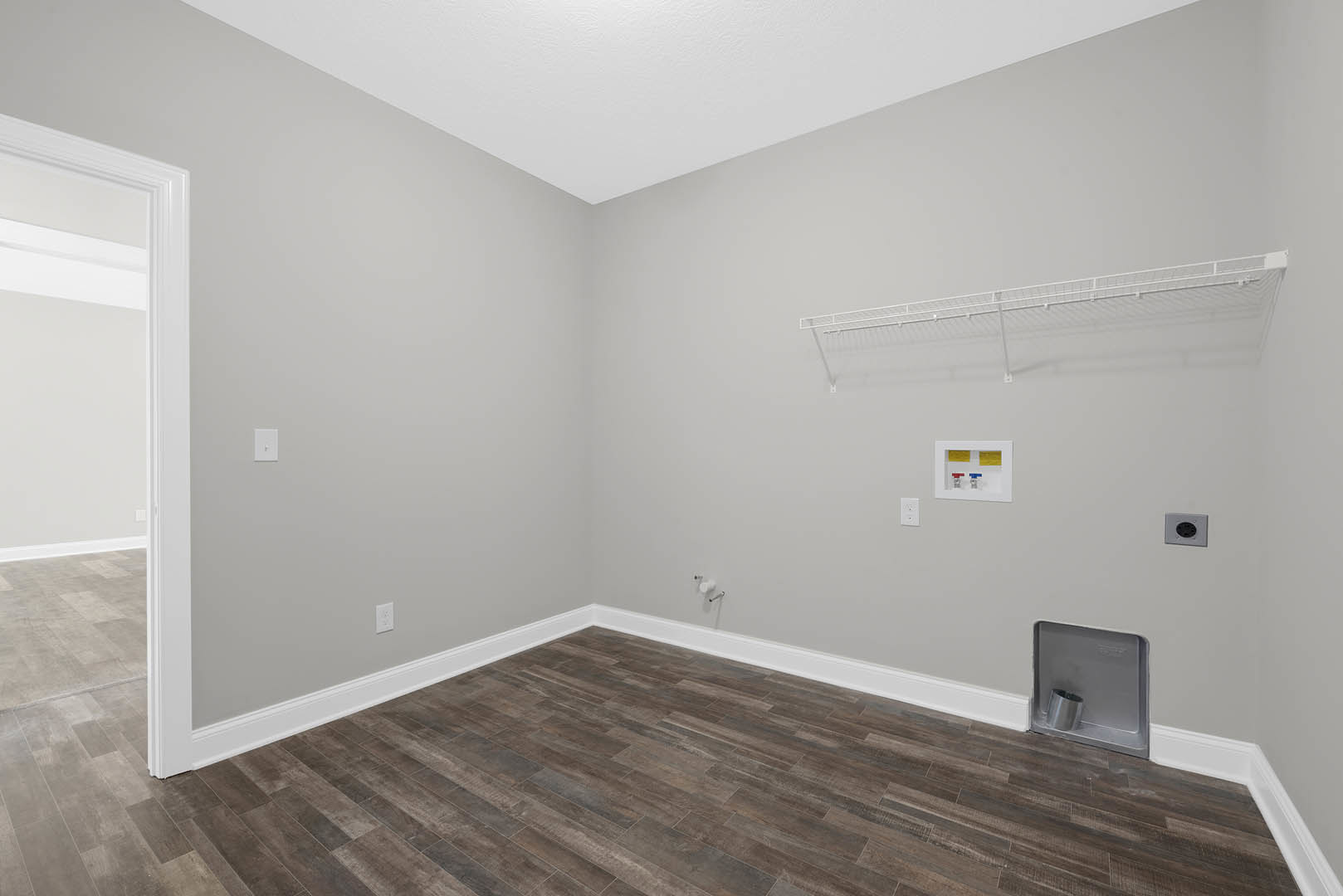 Wood floor with white baseboards, white wall featuring a metal cylinder, white box with red and blue buttons, white shelf with metal shelf, and wall outlet with black circle.