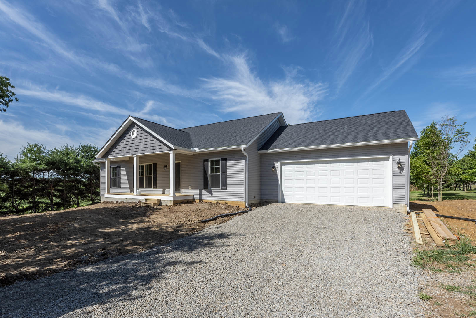 Modern two-story home with gray siding and white trim, attached garage, paved driveway, pile of lumber near the entrance, large windows, surrounded by tall trees under a partly