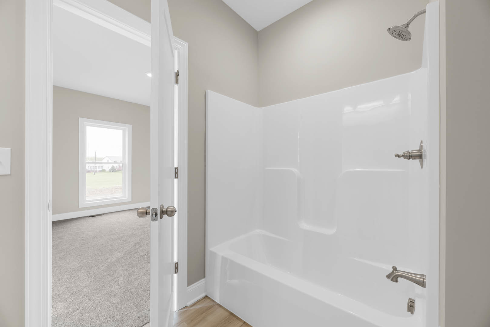 Bathroom with white bathtub and glass-enclosed shower, light tile flooring, chrome shower head, window overlooking neighboring house, white plaster walls, modern door with metal