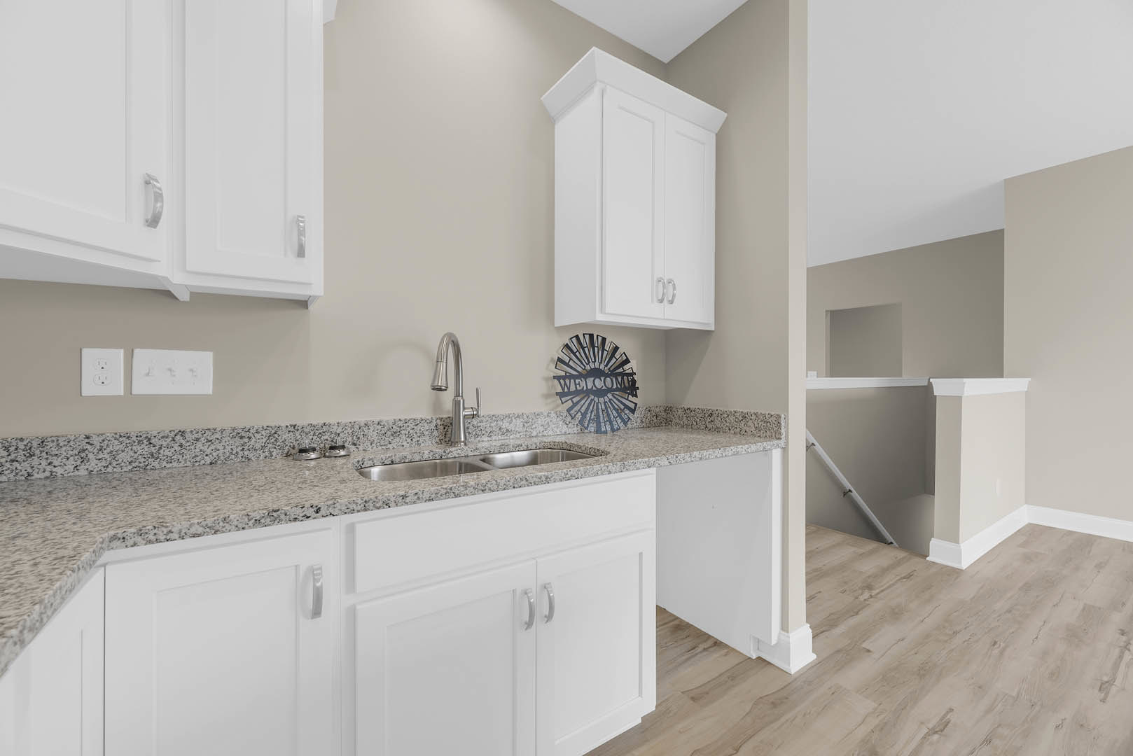 White kitchen with granite countertops, metal welcome sign on wall, white cabinetry with corner detail, stainless steel faucet and sink, white railing along wall