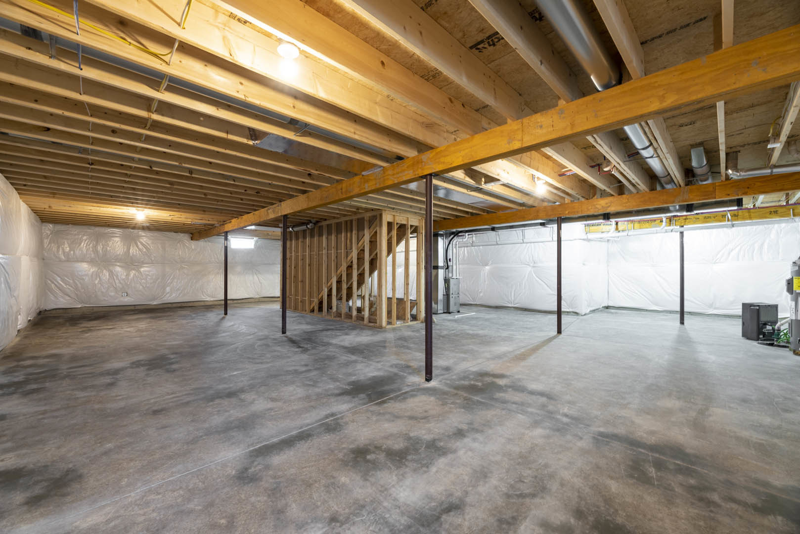Basement room featuring exposed wooden ceiling beams, white wall partially covered with plastic sheeting, concrete floor, open black utility box, and staircase leading upward.