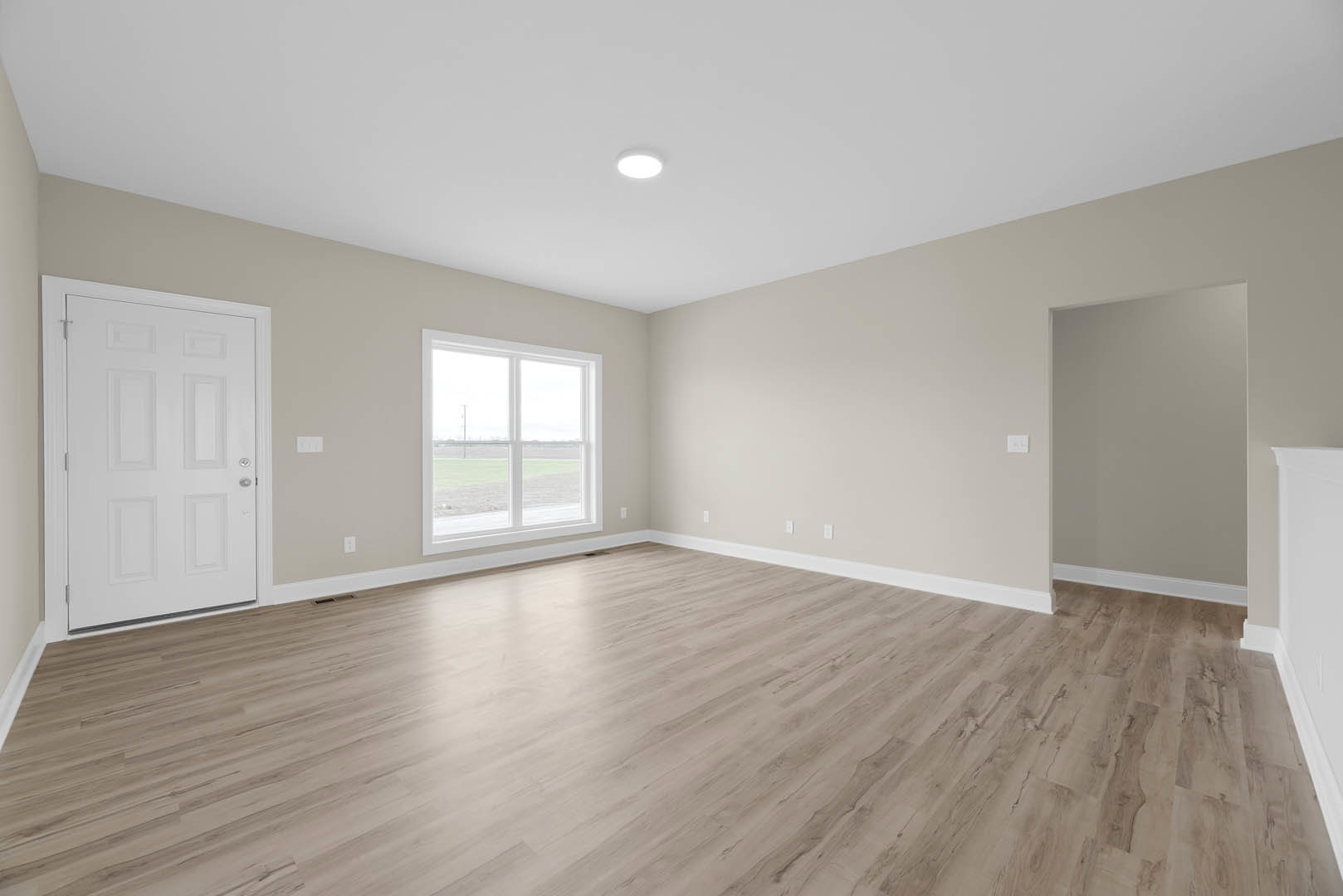 Room with light wood flooring, white walls with black trim, white door featuring a silver handle, ceiling light fixture, and window overlooking a grassy field.