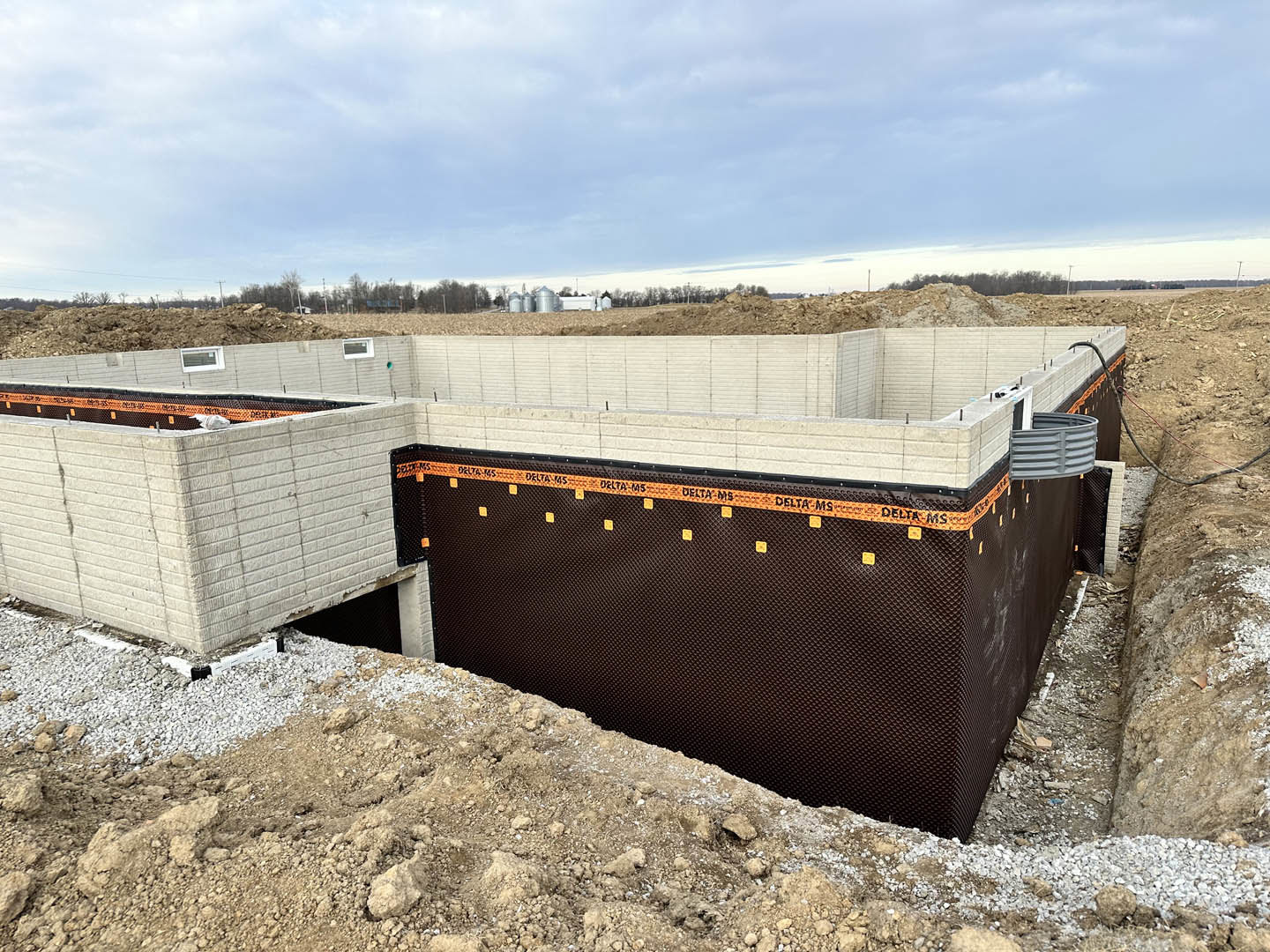 Framed exterior walls with exposed composite materials, construction debris and dirt in foreground, blue sky with scattered clouds above