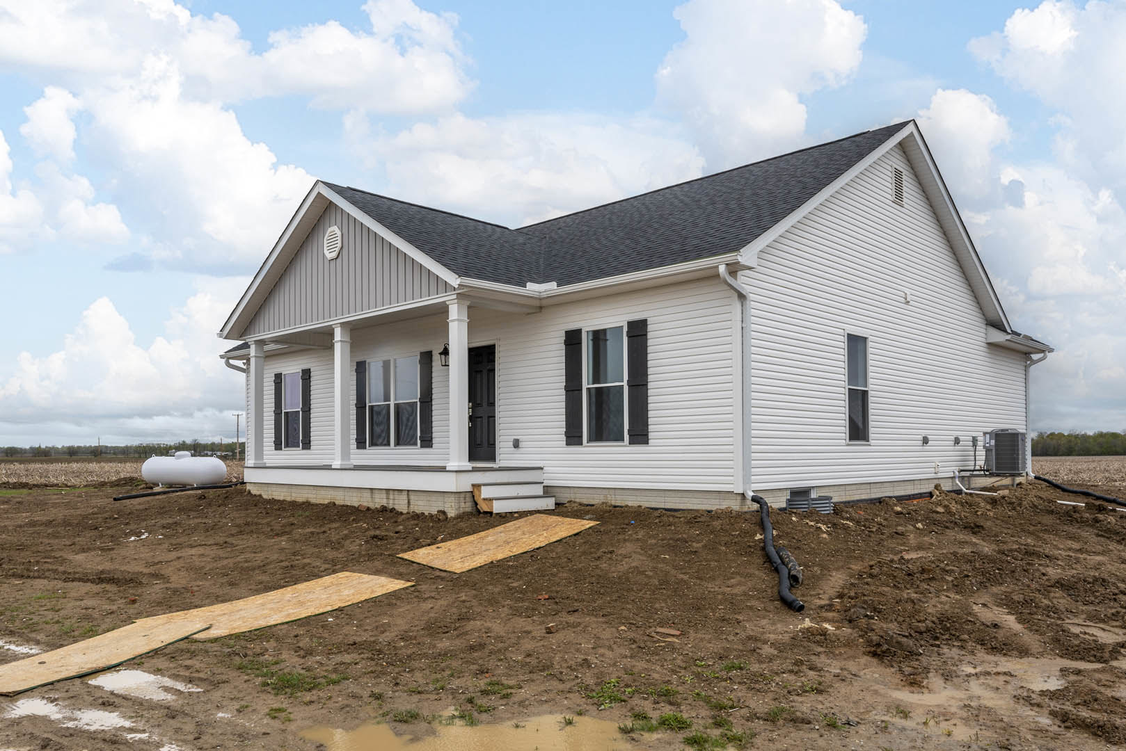 White house with black shutters and black roof under construction, dirt ground with scattered plywood and black pipe, blue sky with clouds in background