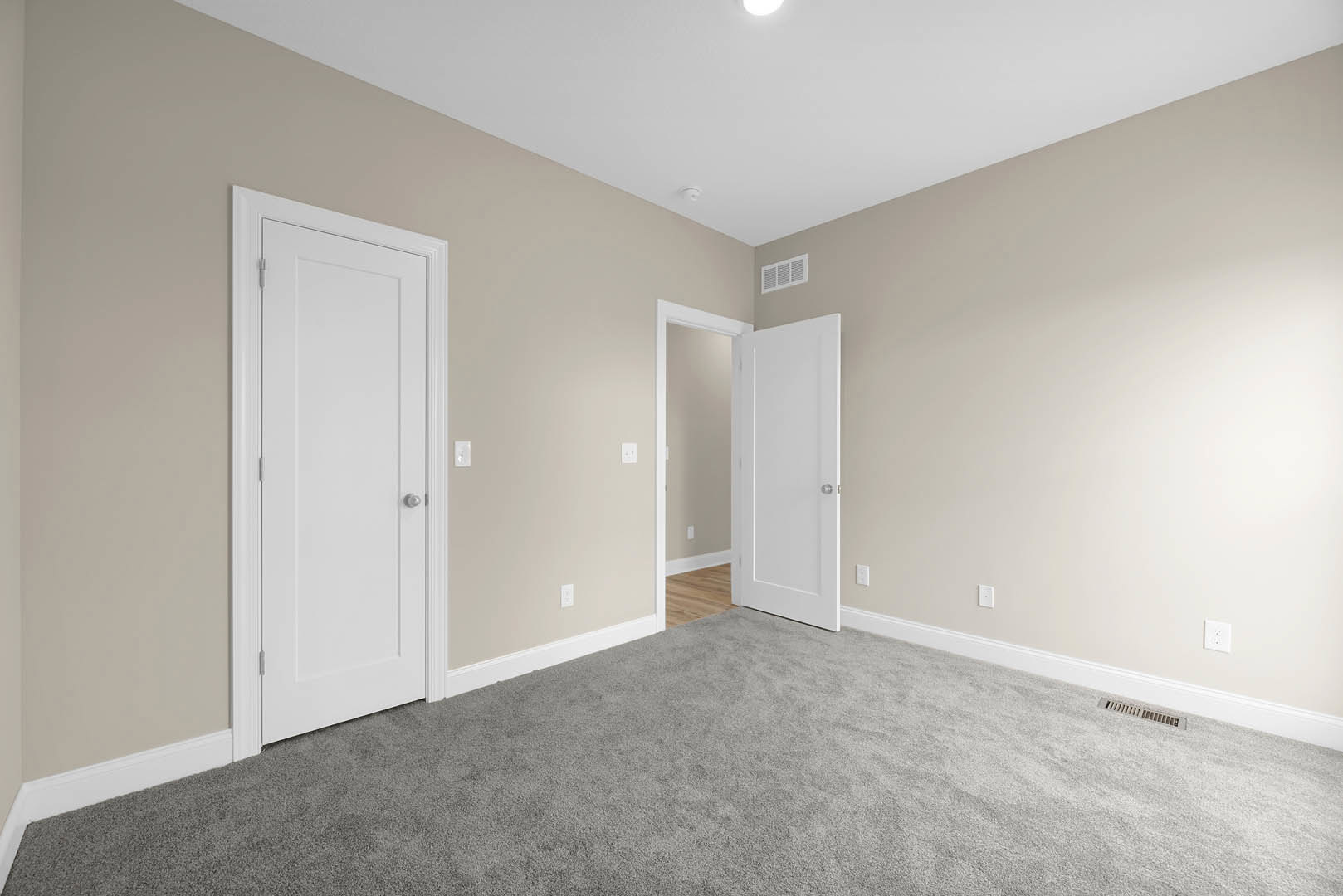 Carpeted room with two white doors featuring silver knobs, white walls with base molding, and a wall vent