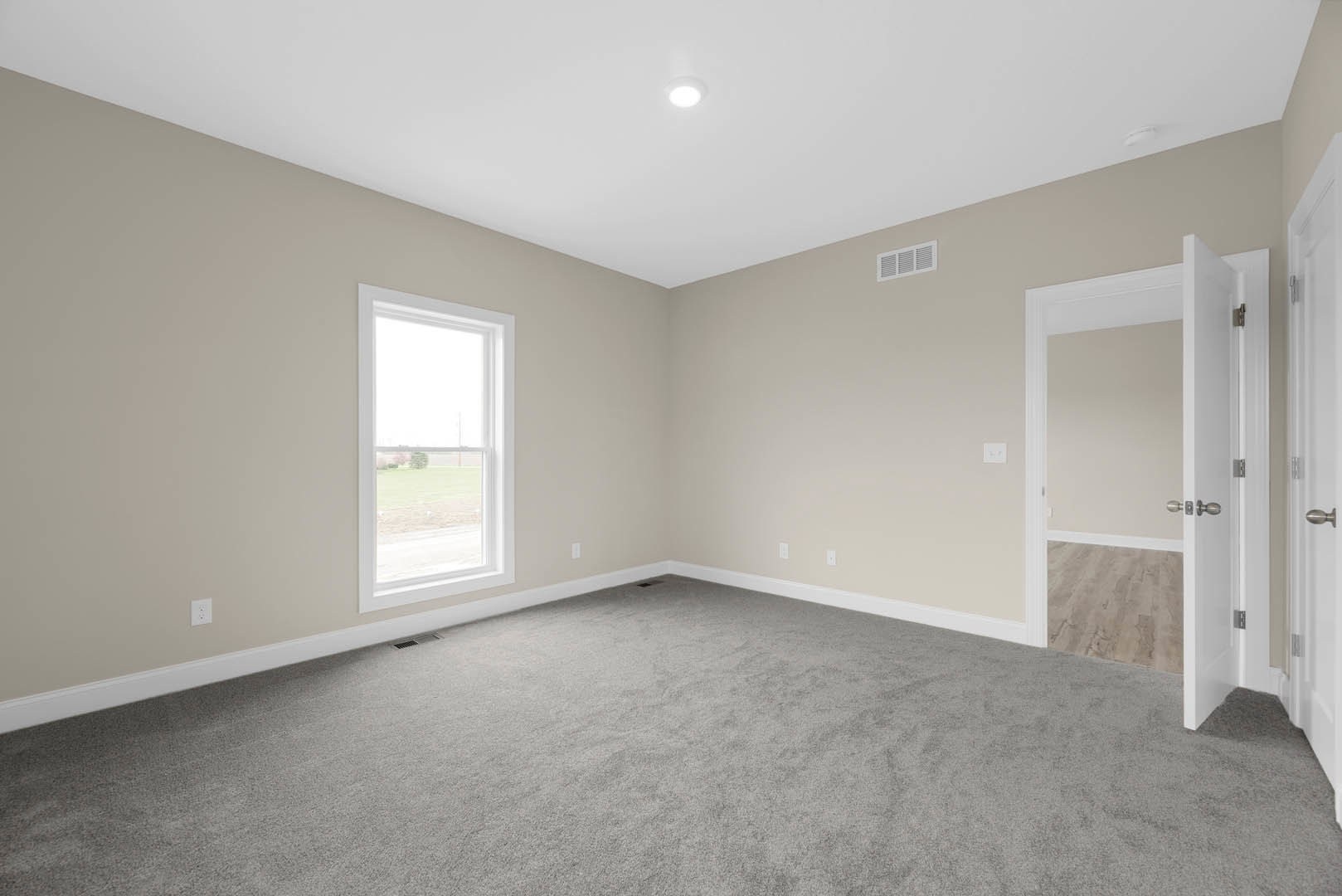 Carpeted room with white walls, large window overlooking grassy field, ceiling light fixture, wall vent, and white trim.
