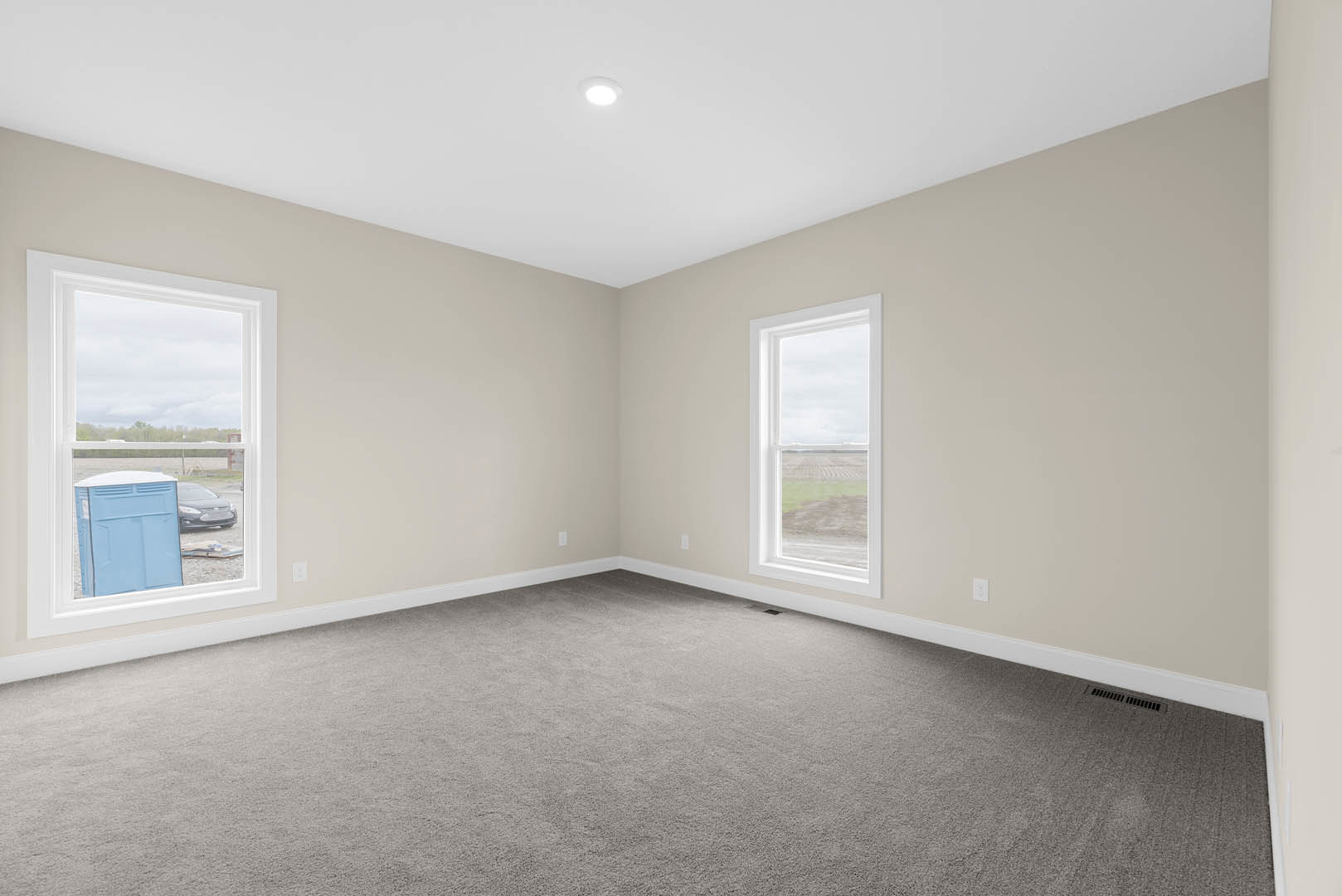 Carpeted room with white walls, multiple windows, ceiling light fixture, and view of a grassy field and blue portable toilet outside.