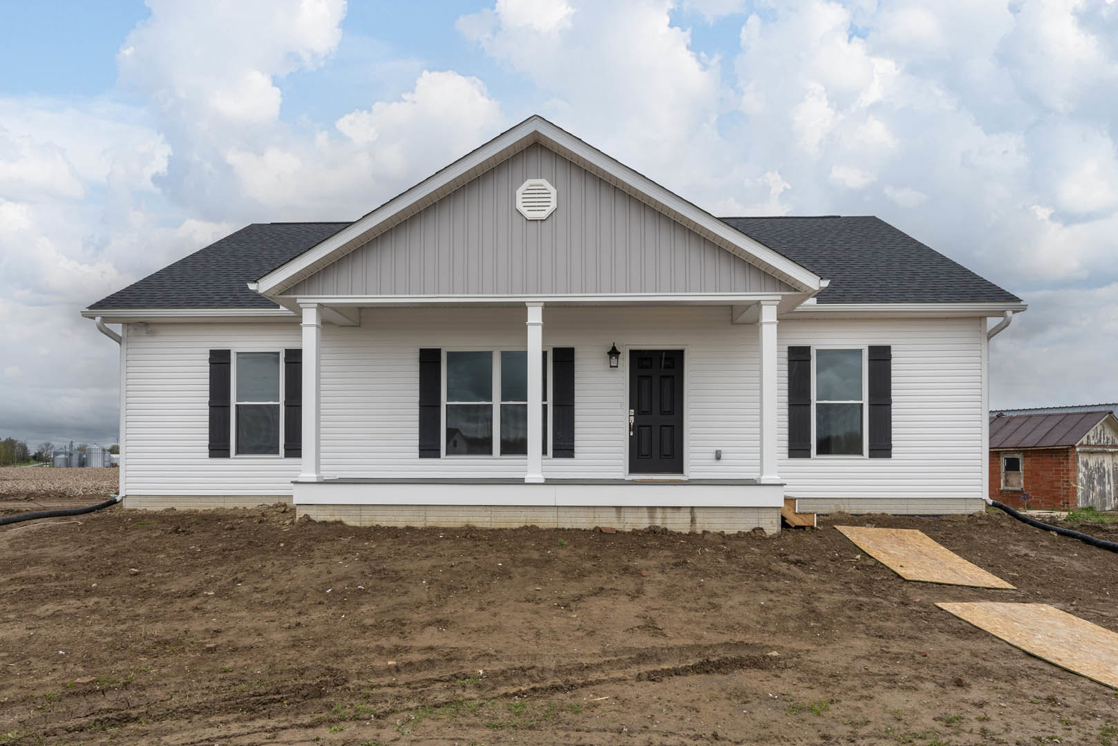 White siding house with black shingle roof, black front door framed in white, white vent on gray wall, dirt patch with tire tracks in foreground, large windows, cloudy sky