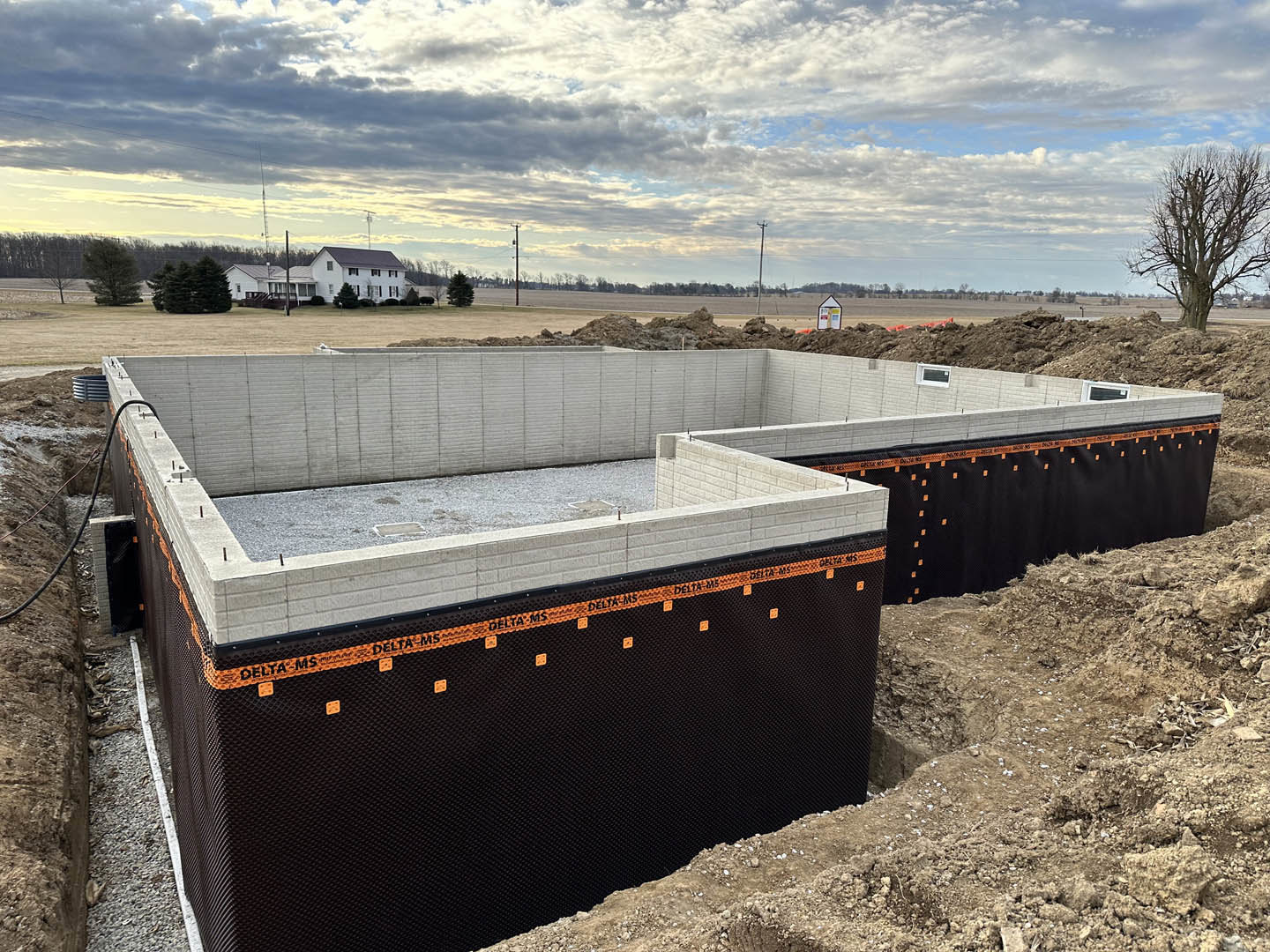 Partially built concrete foundation surrounded by gravel and dirt, leafless trees and cloudy sky in the background, black fence along construction site perimeter