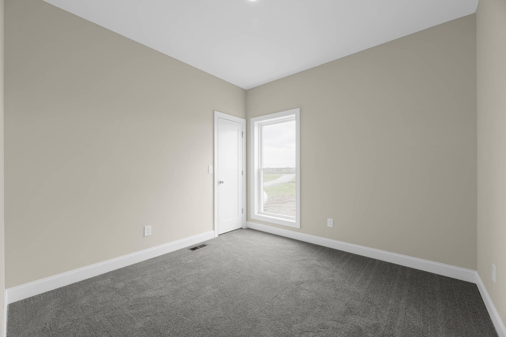 Grey carpeted bedroom with white walls, white ceiling light fixture, white door featuring silver knobs, and window overlooking grassy yard