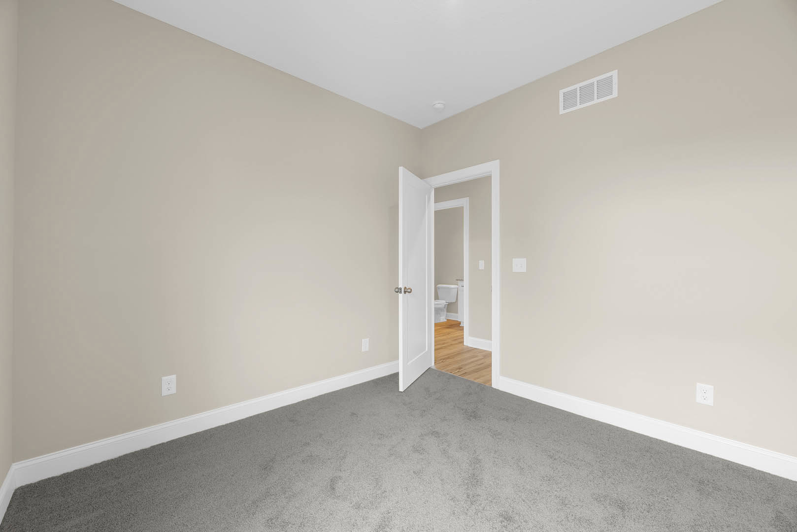 Open white door leading from a grey carpeted bedroom to a bathroom with wood flooring, white toilet, plastic container, and ceiling vent